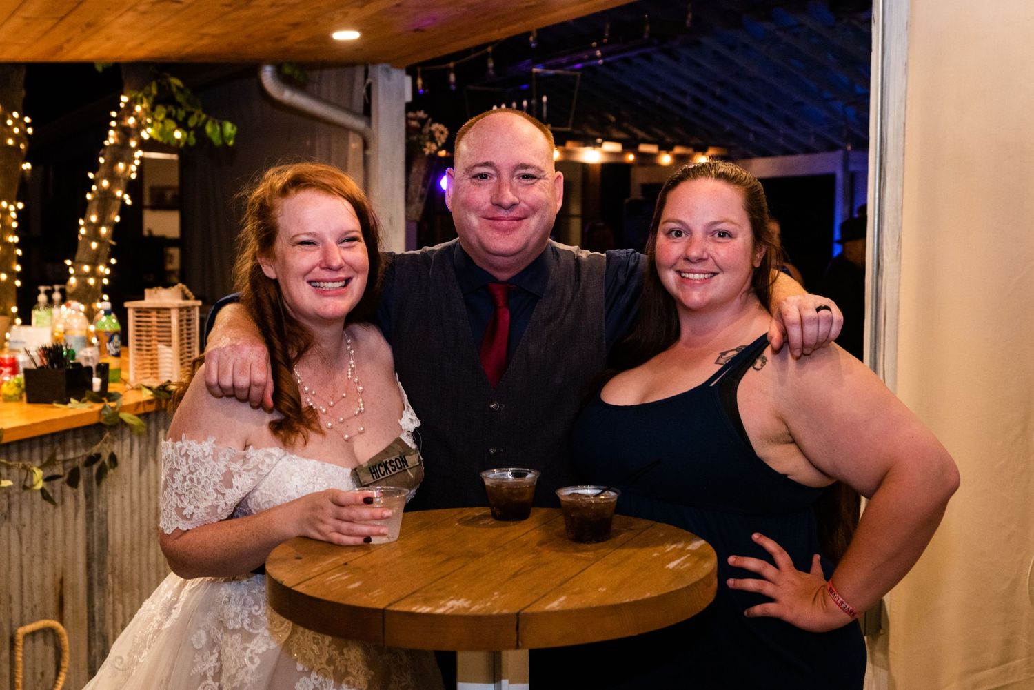 Wedding guests pose together at a wooden high table during an evening reception celebration.