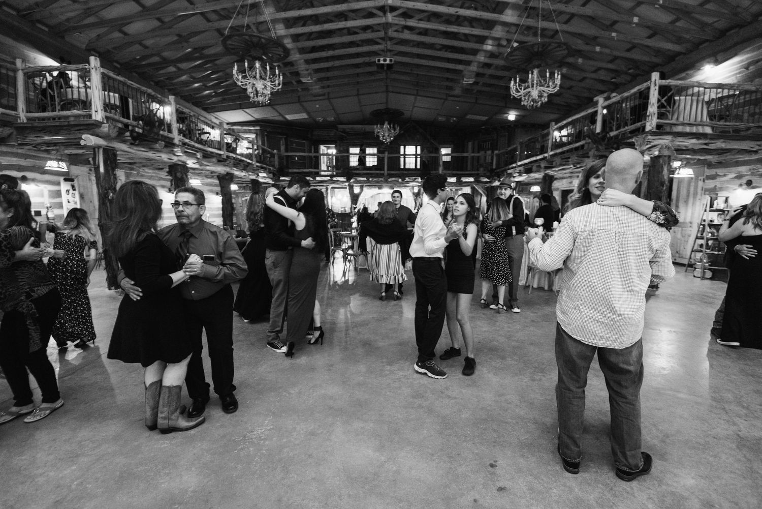 Black and white photo of couples dancing at a rustic barn venue with chandeliers overhead.