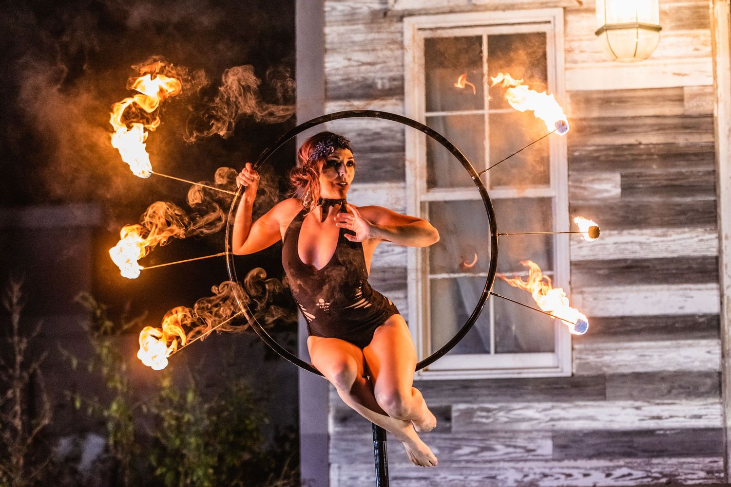 Aerial artist on circular prop performs fire dance routine against weathered wooden building.