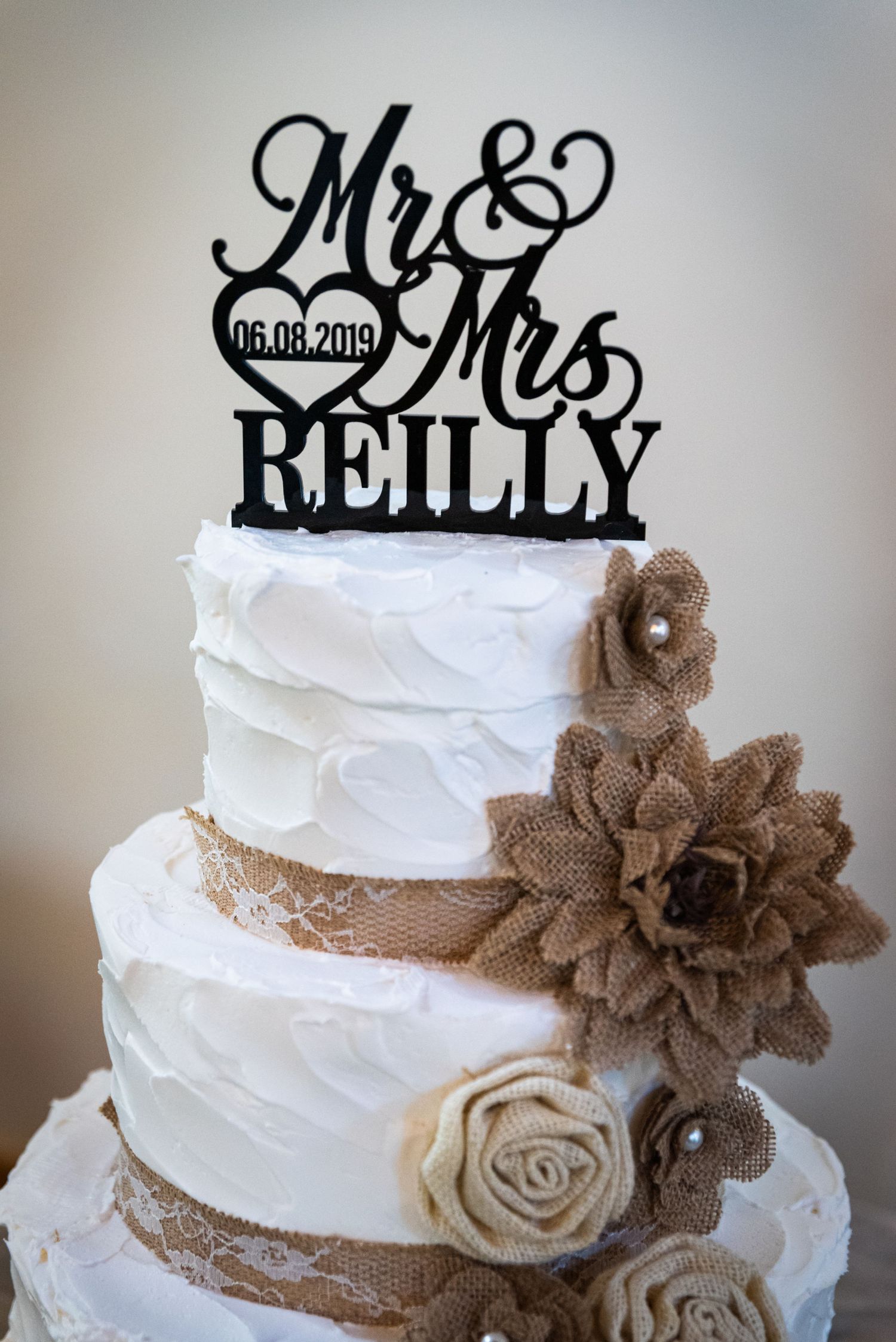 White wedding cake with 'Mr & Mrs Reilly' topper and burlap flower decorations.