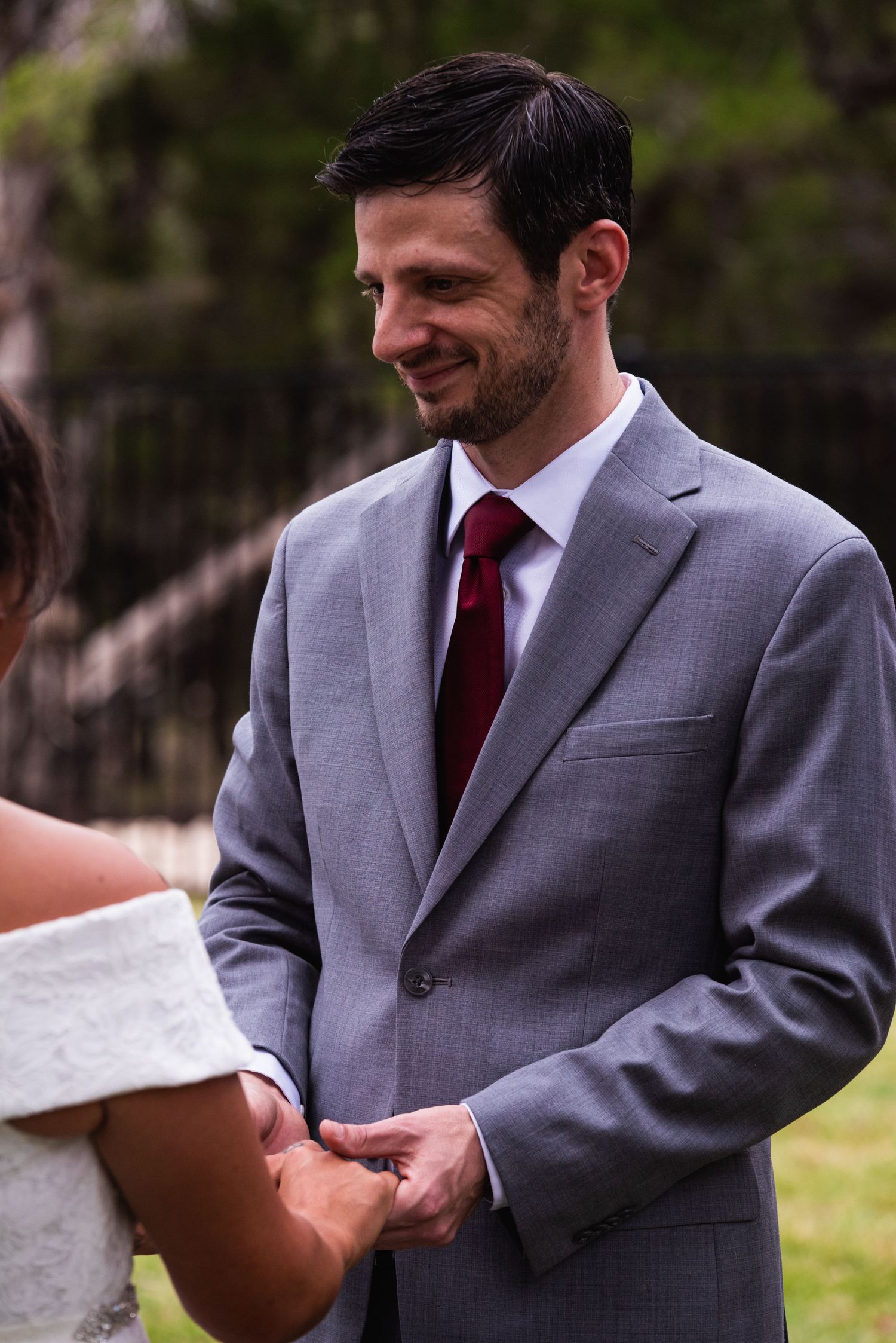 Groom in grey suit and red tie smiles warmly during outdoor wedding ceremony.