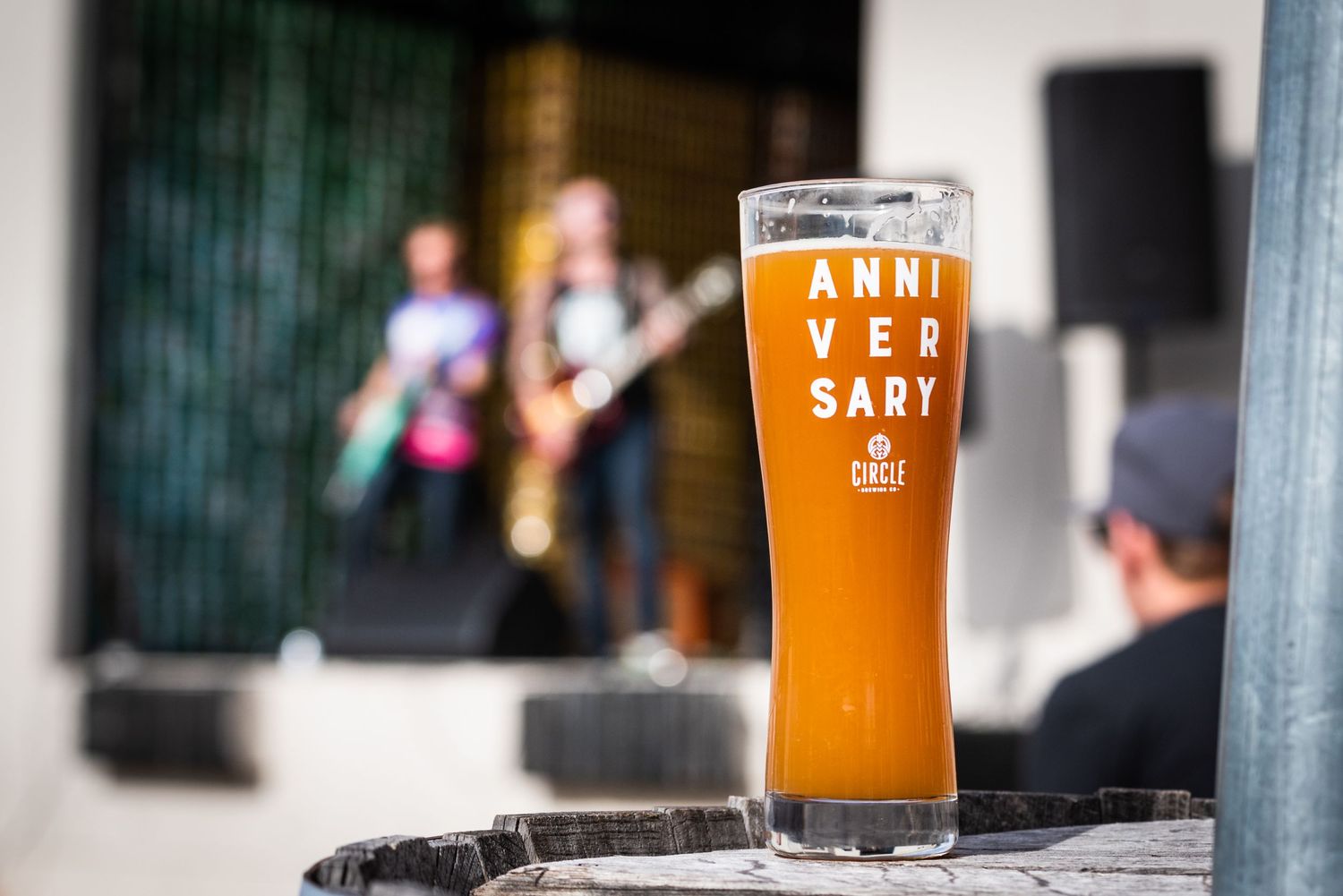 A tall glass of golden beer with 'Anniversary' text sitting on a ledge with blurred background.