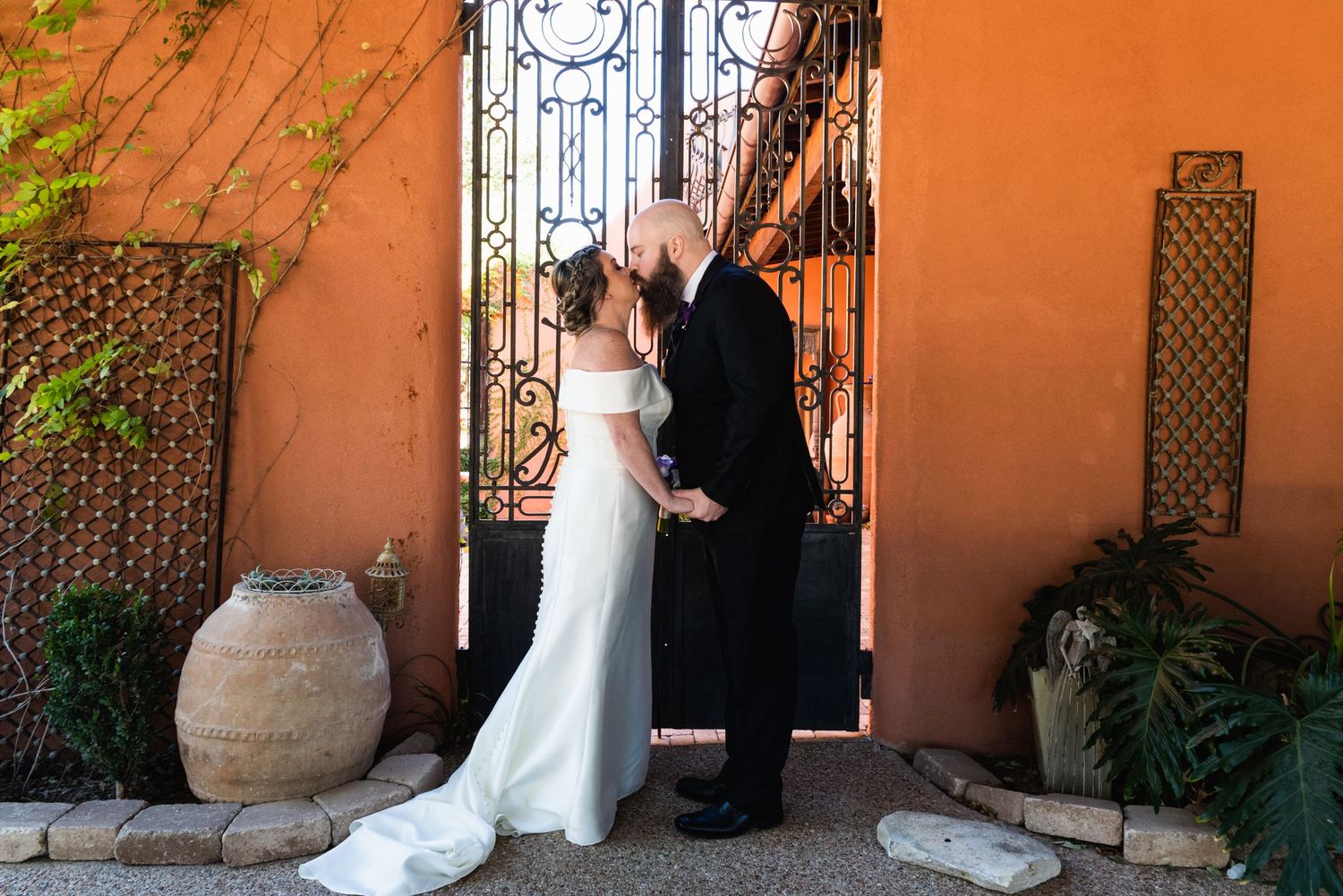 Couple shares intimate moment by ornate iron gate against orange stucco wall with decorative pottery.