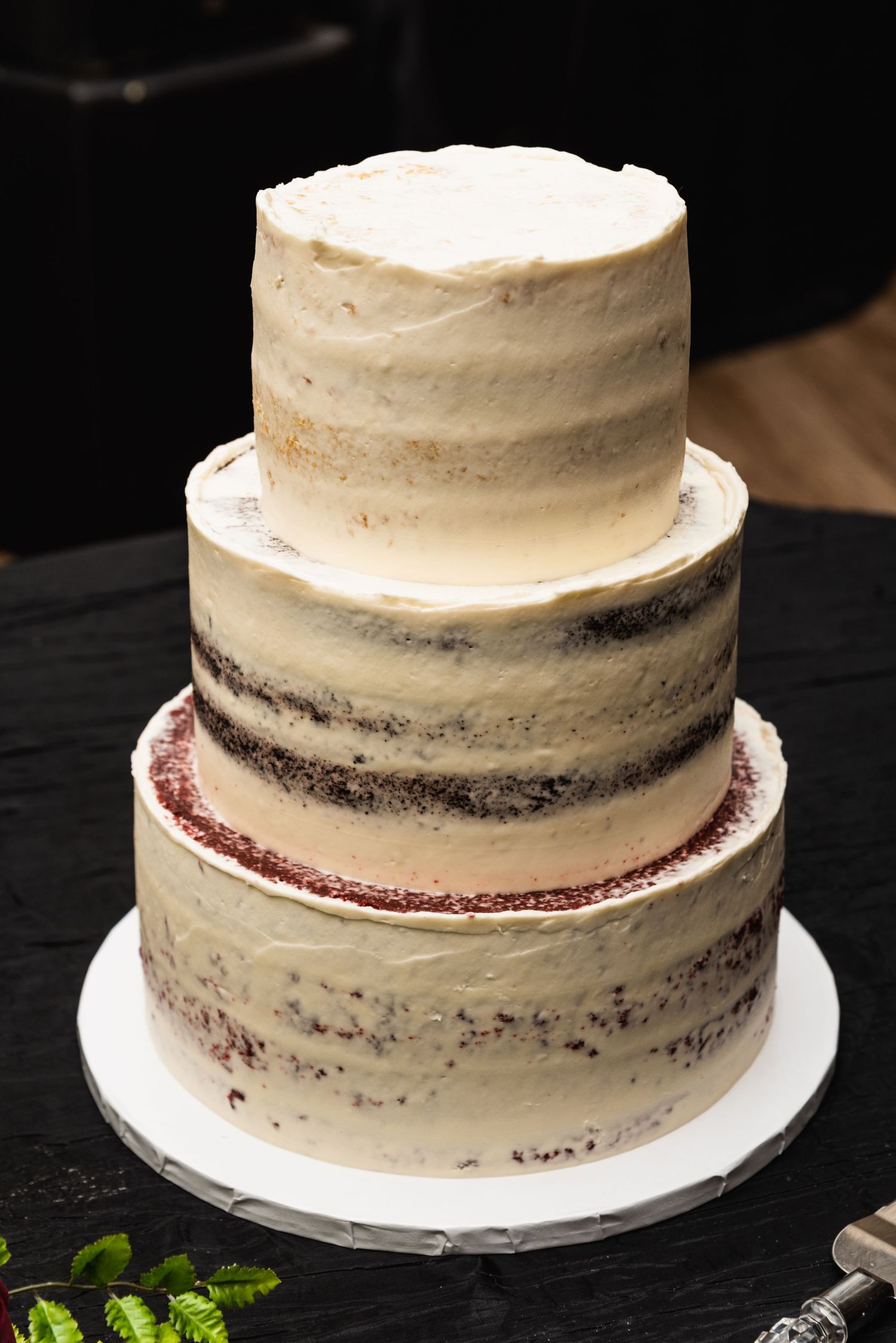 A three-tier semi-naked wedding cake with textured white frosting on a black tablecloth.