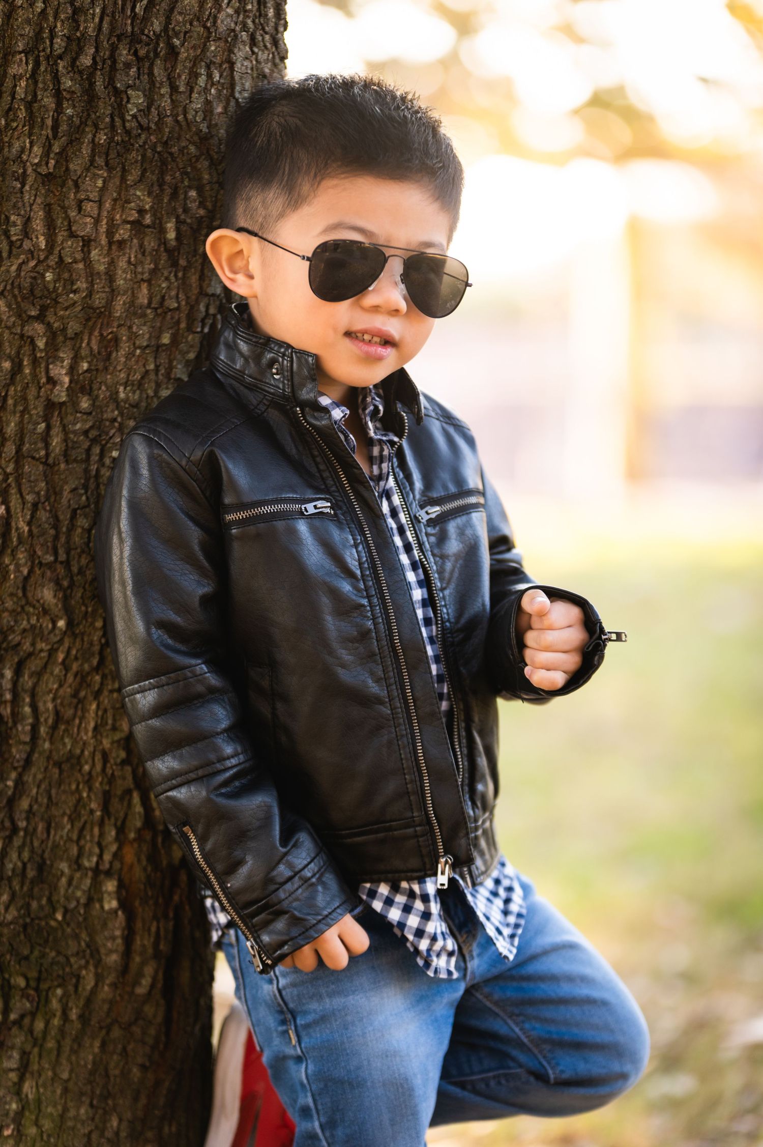 A stylish child poses against a tree wearing sunglasses and a leather jacket.