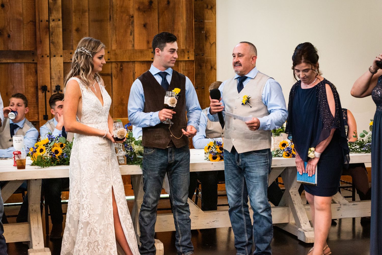Wedding party members share toasts and conversation during reception in rustic venue setting.