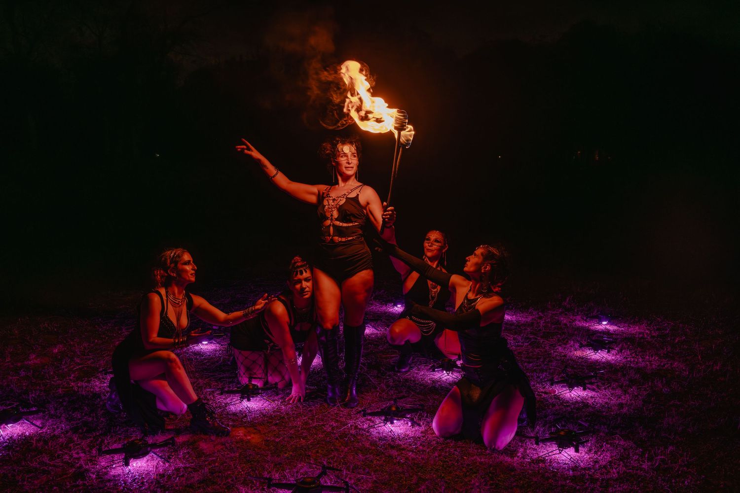 Fire dancer performs with flaming torch surrounded by women kneeling in purple light at night outdoor show.