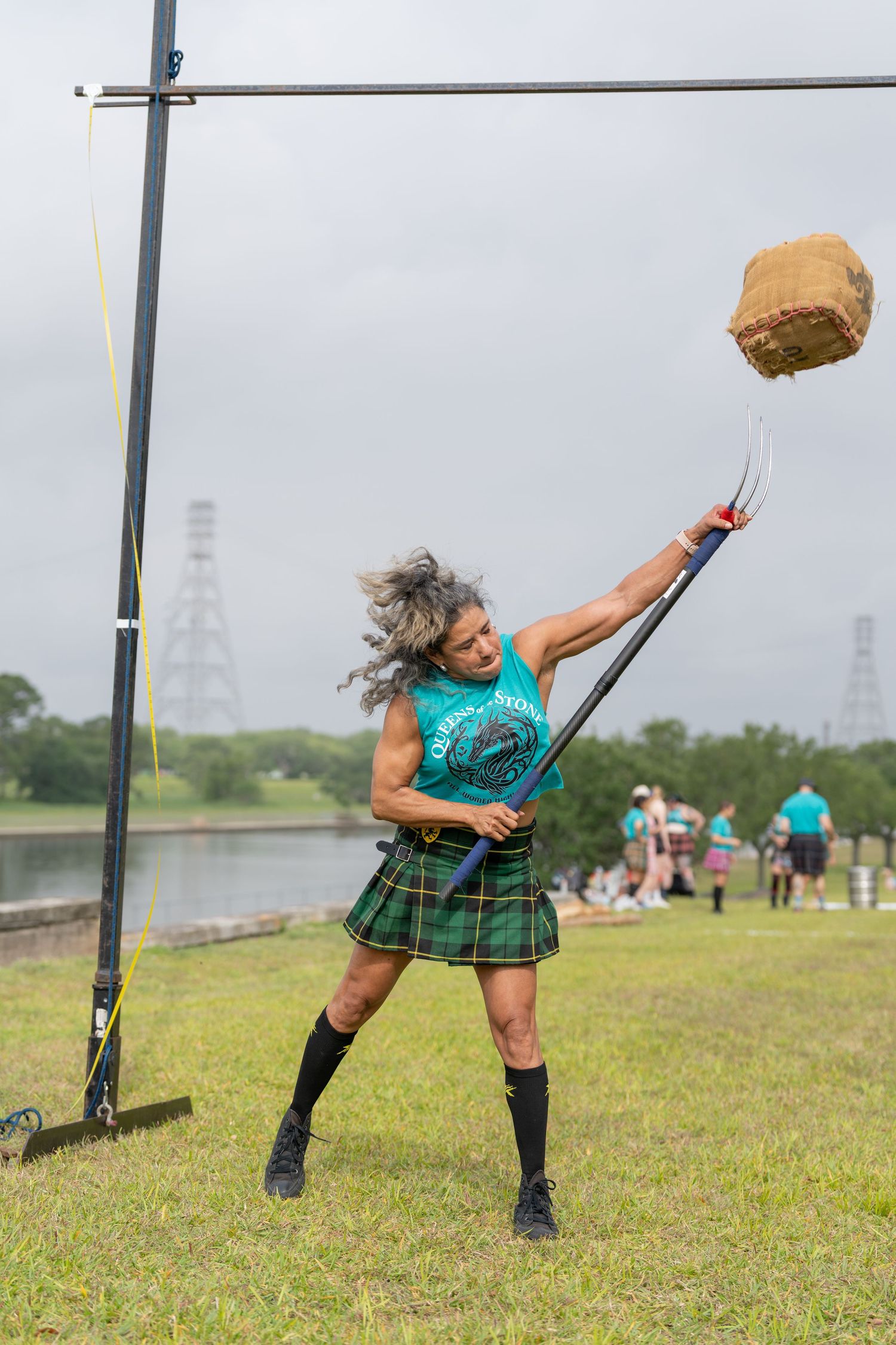 Woman in tartan kilt competes in Highland Games weight over bar throw on cloudy day.