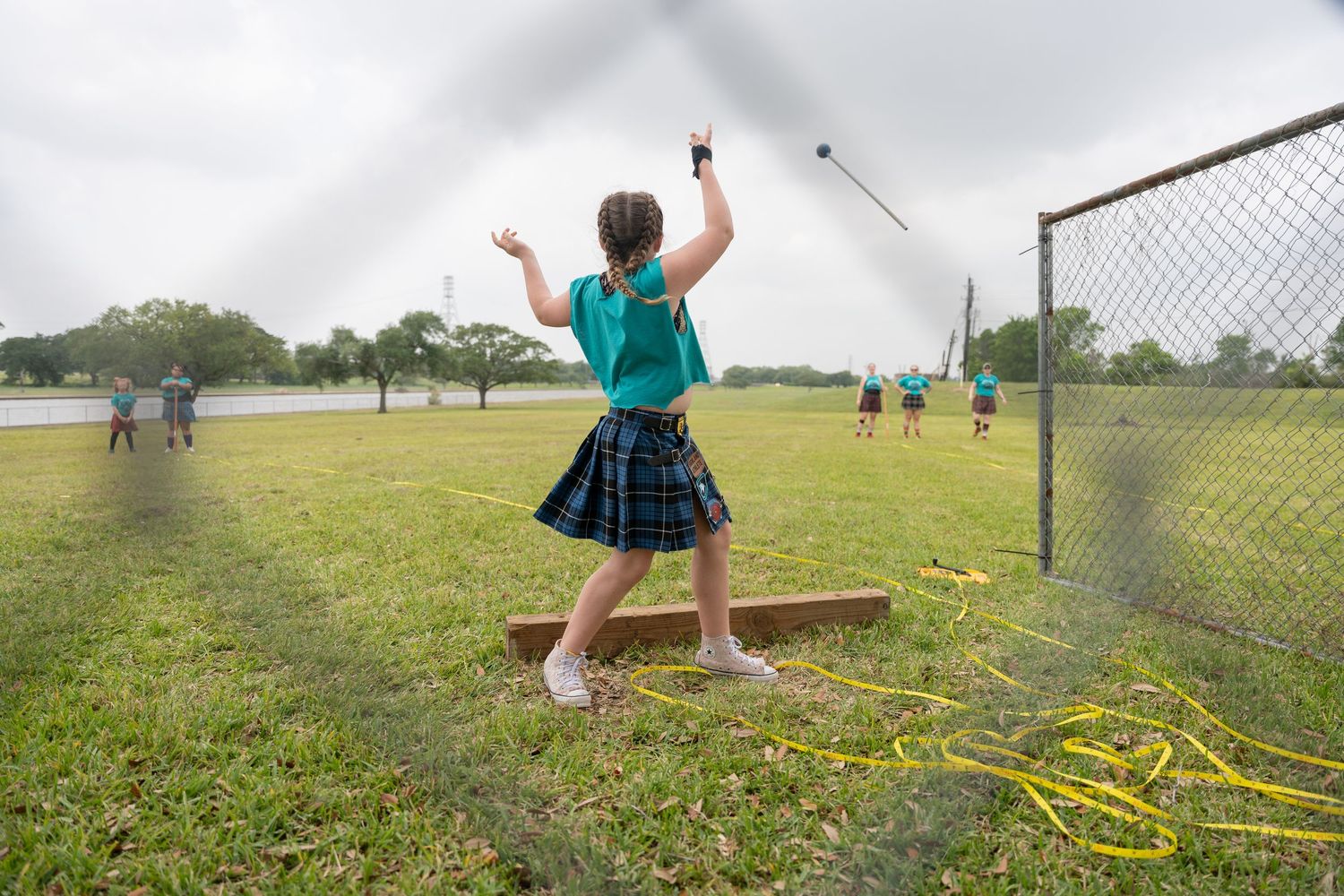 Girl in plaid skirt throwing a javelin on a grassy field during an outdoor track and field event.