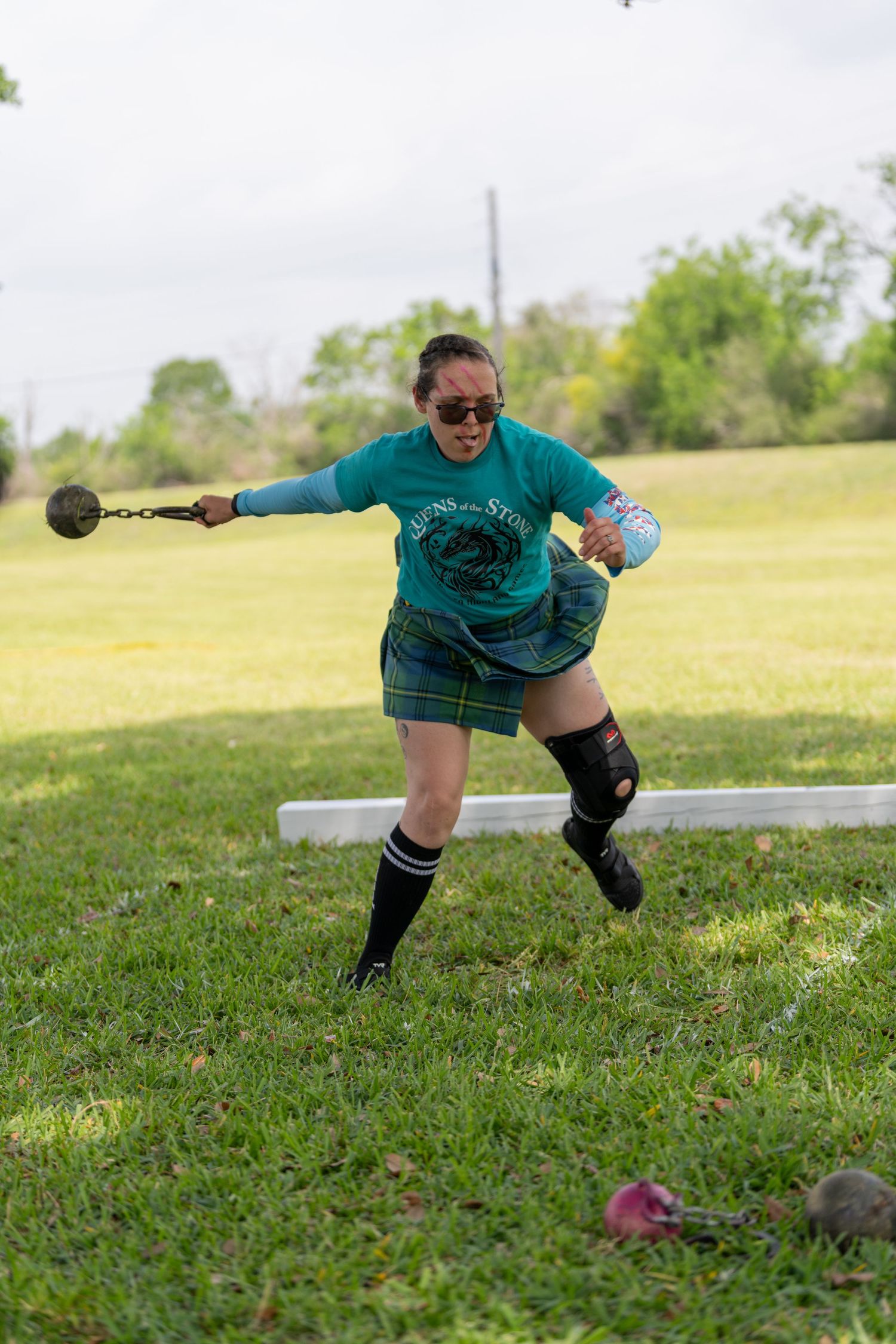 Woman in green shirt and plaid kilt throws a weight on a chain during a Highland Games competition outdoors.