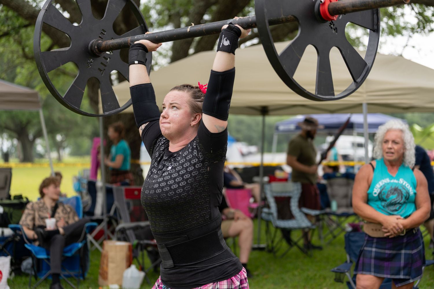 Woman lifting heavy barbell overhead at outdoor Highland Games competition with spectators watching in background.