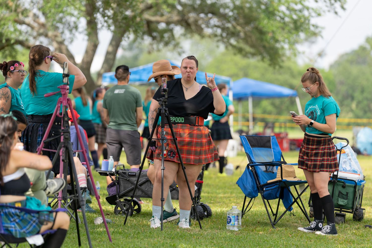 People in kilts and teal shirts gather at an outdoor Scottish Highland Games festival with cameras and folding chairs.