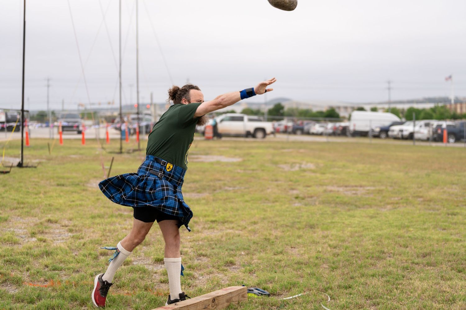 Athlete in blue tartan kilt throws heavy ball at Highland Games competition on green field.