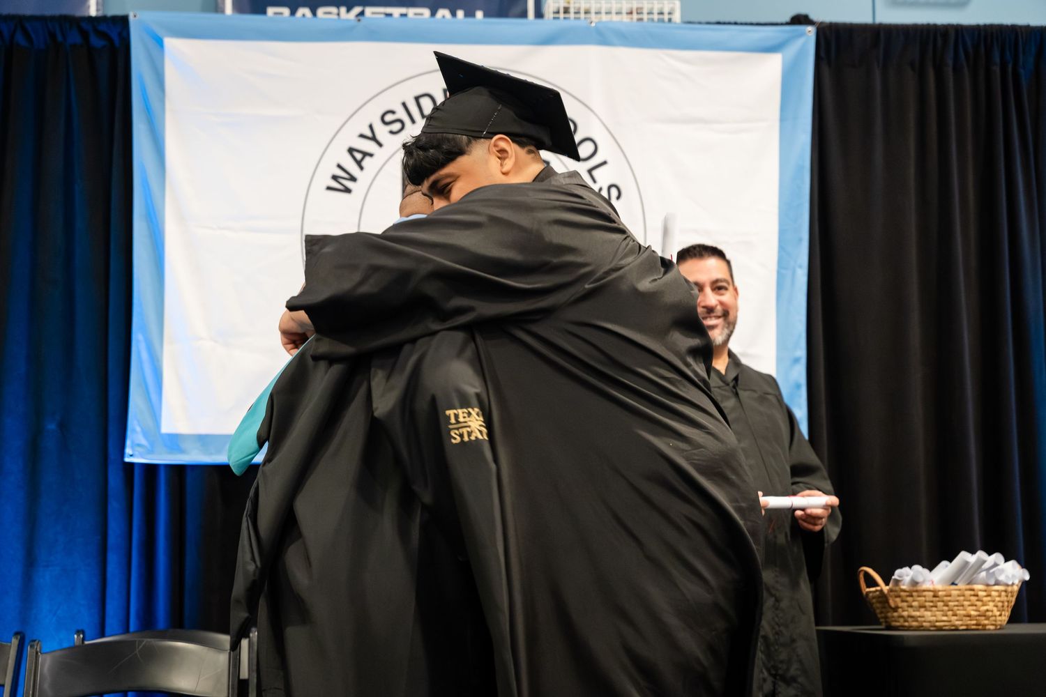 Two graduates in black robes embrace at a Wayside School graduation ceremony, with a smiling man standing nearby.