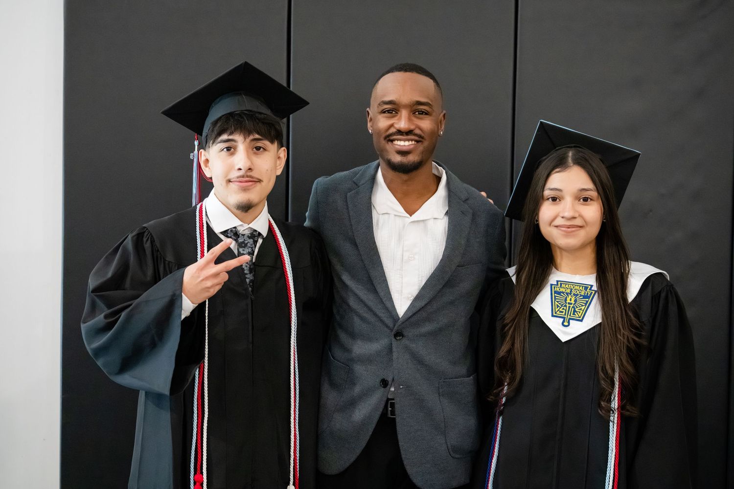 Two graduates in black caps and gowns pose with a man in a suit at a graduation ceremony.