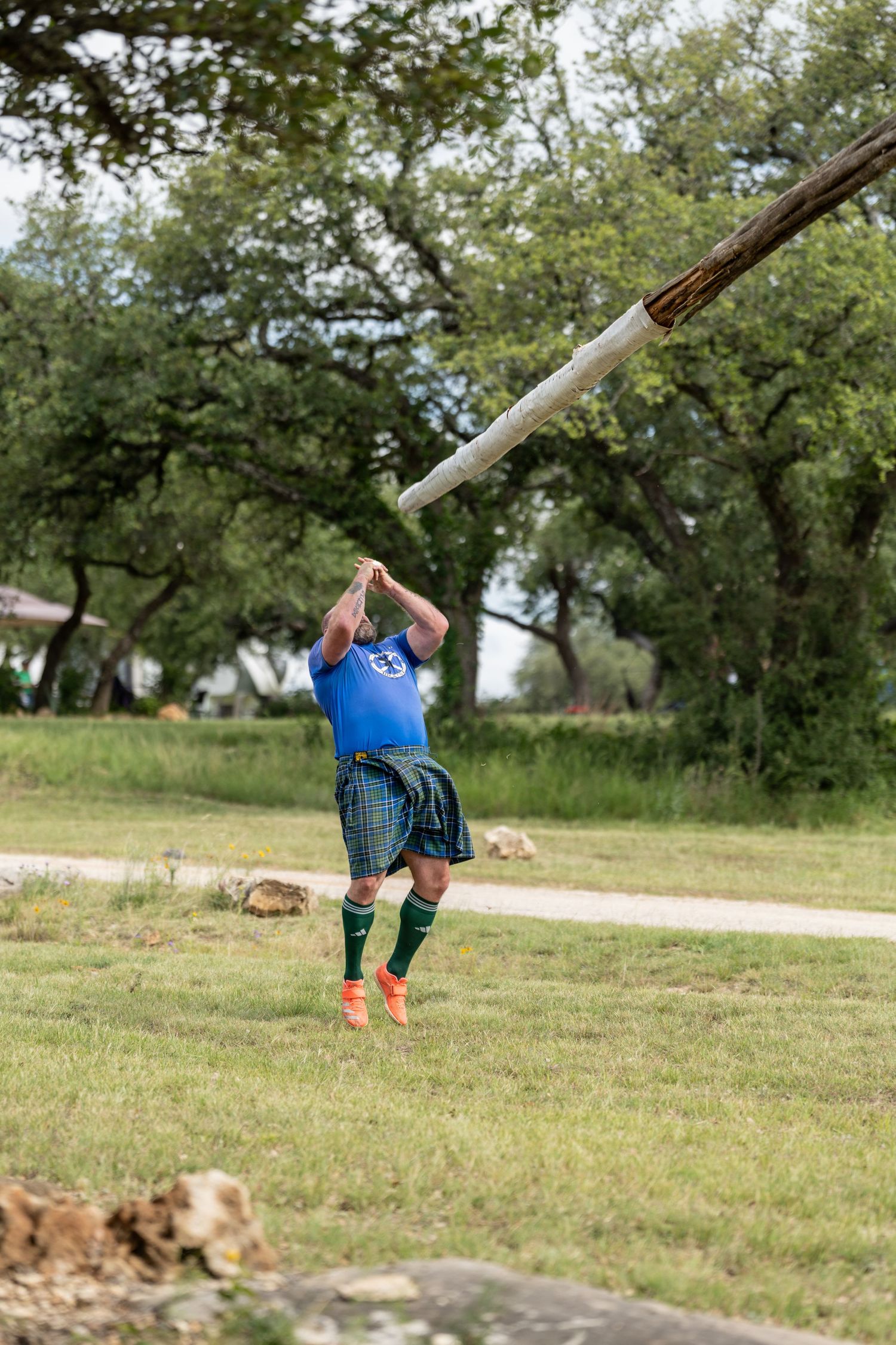 Man in blue shirt and tartan kilt tossing a caber at a Highland Games competition outdoors.