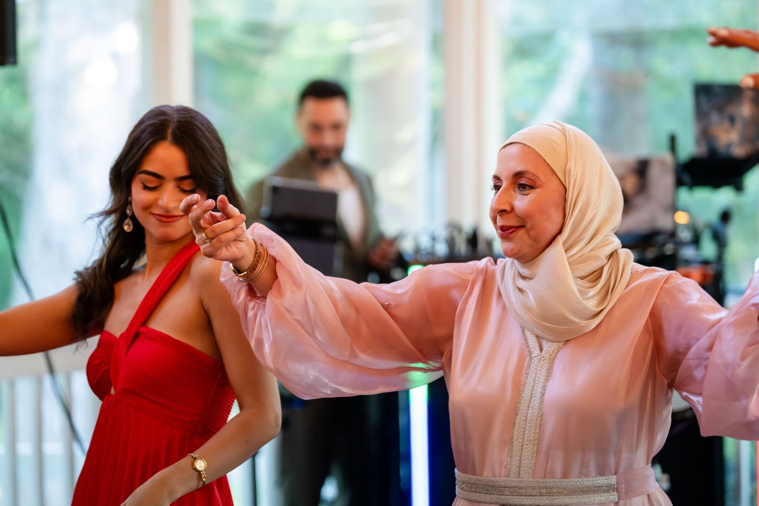 Two women in elegant attire dance together at a celebration, one in red dress, other in pink hijab.