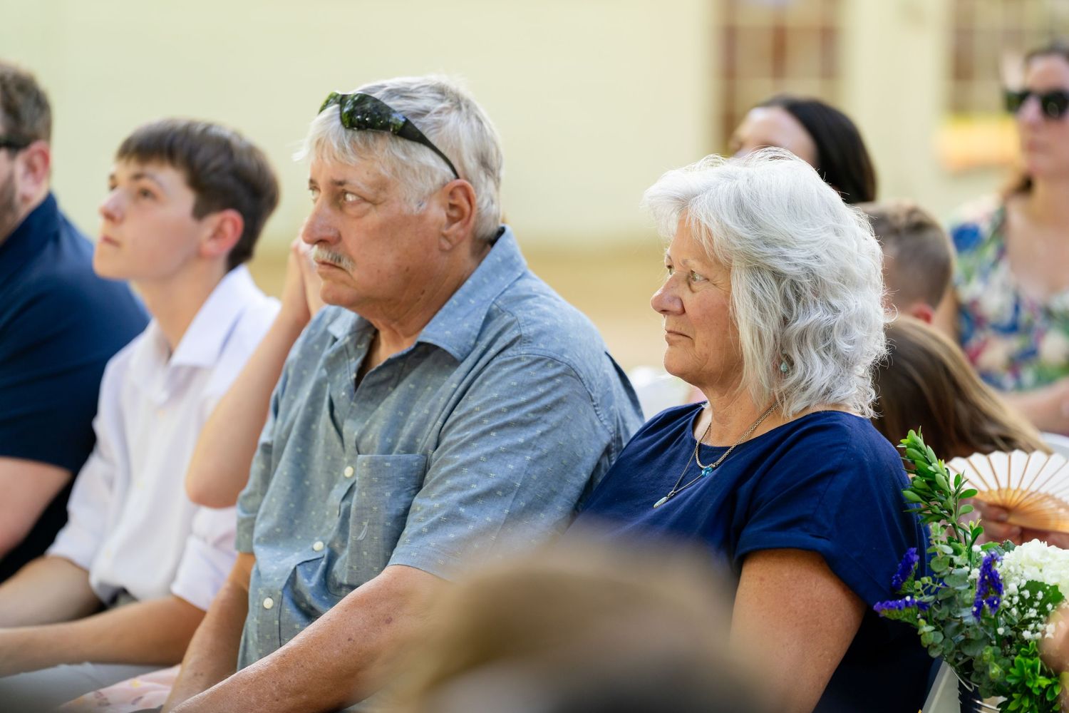 Elderly couple sitting attentively outdoors at a sunny event, woman holding a decorative fan, people seated in background.