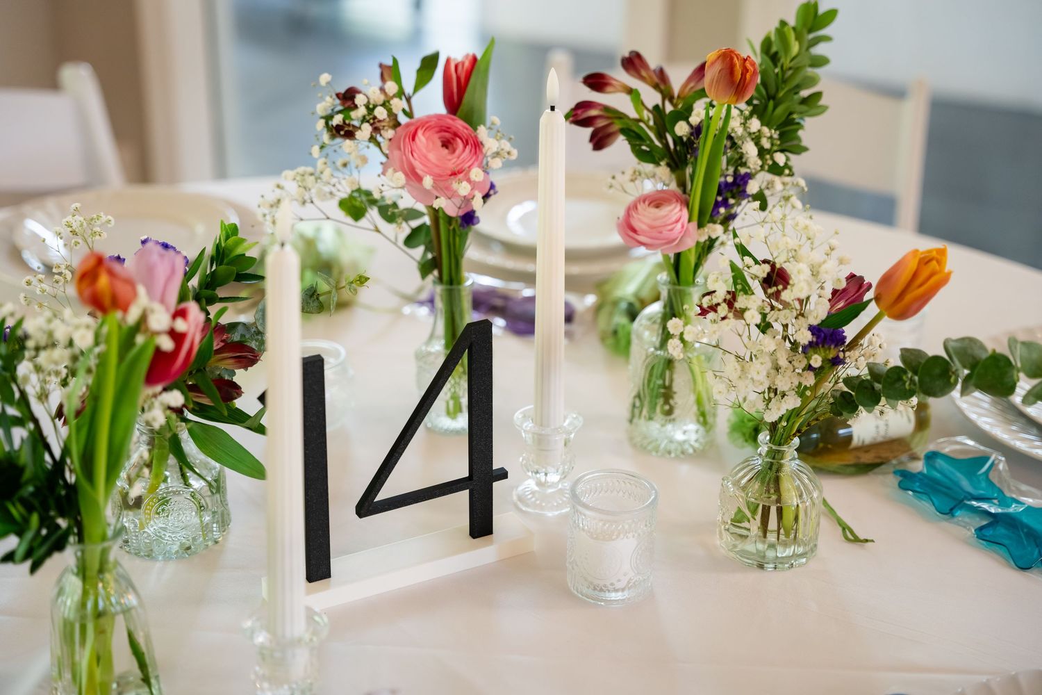 Wedding table centerpiece with colorful tulips, ranunculus, and wildflowers in glass vases beside a table number 14 sign.