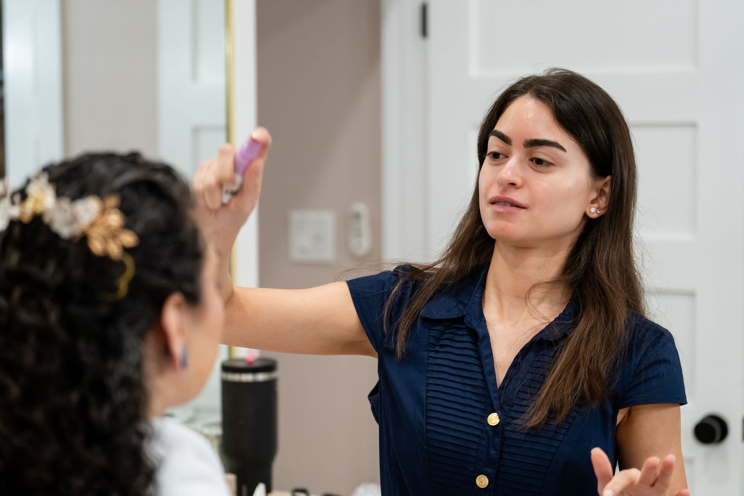 A woman in a navy blue top applies makeup or demonstrates a beauty technique on another person in a bright room.