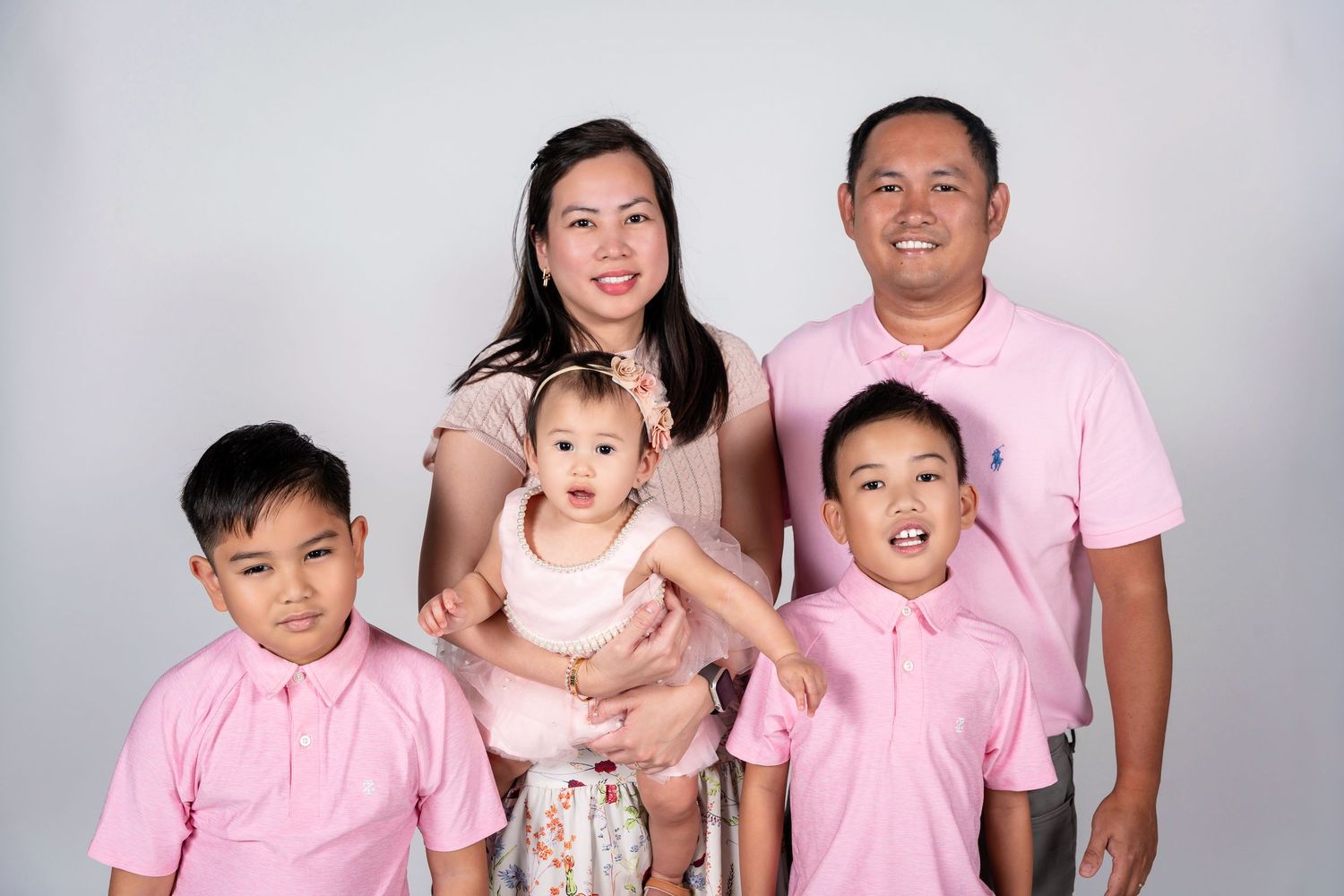 Family of five in matching pink outfits posing for a studio portrait, parents holding baby girl with two young boys.