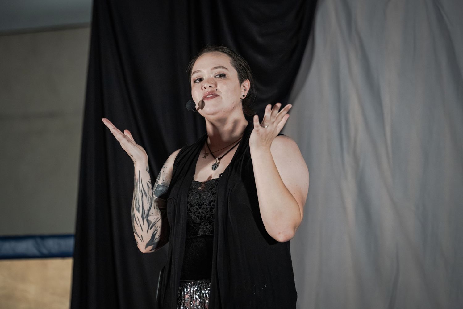 Woman in black vest gesturing expressively on stage with tattoo sleeve, performing in front of dark curtain.