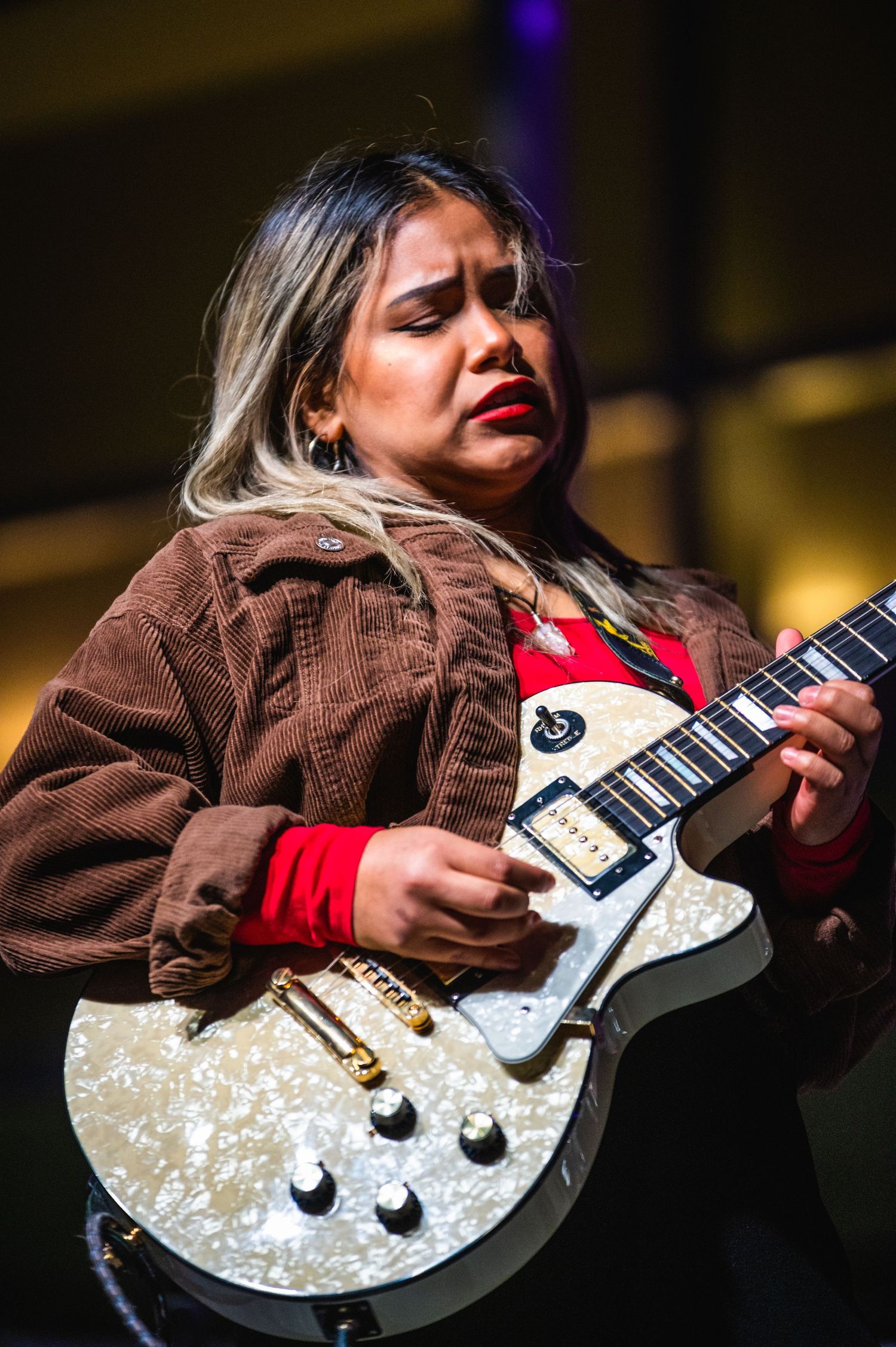 A musician in a brown jacket plays a white electric guitar with dramatic lighting and dynamic performance angles.