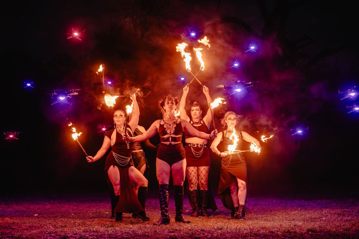 Fire performers pose dramatically at night, wielding flaming torches with colorful lights glowing in the dark background.