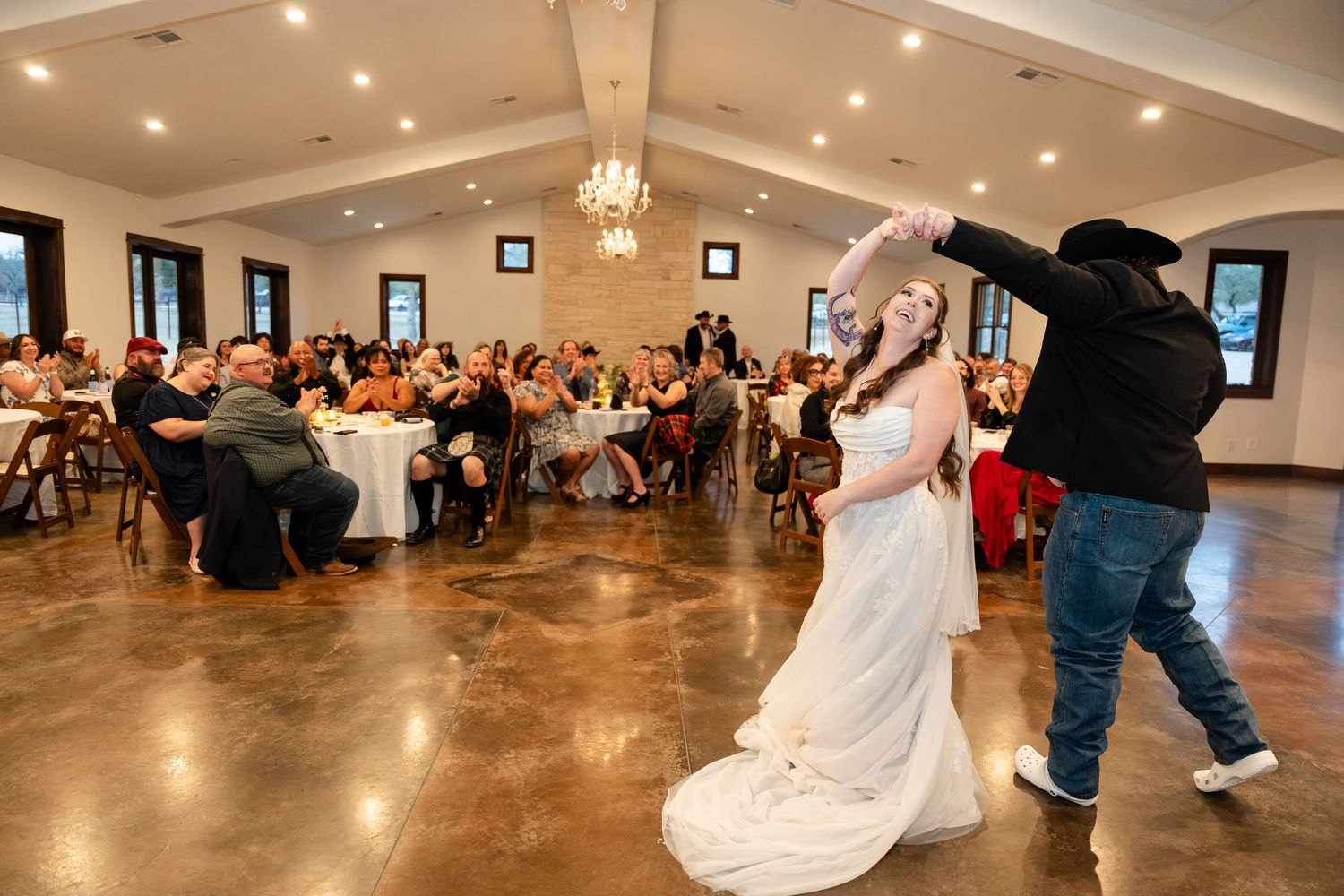 Bride and groom share first dance at rustic wedding reception hall with guests seated at decorated tables in background.