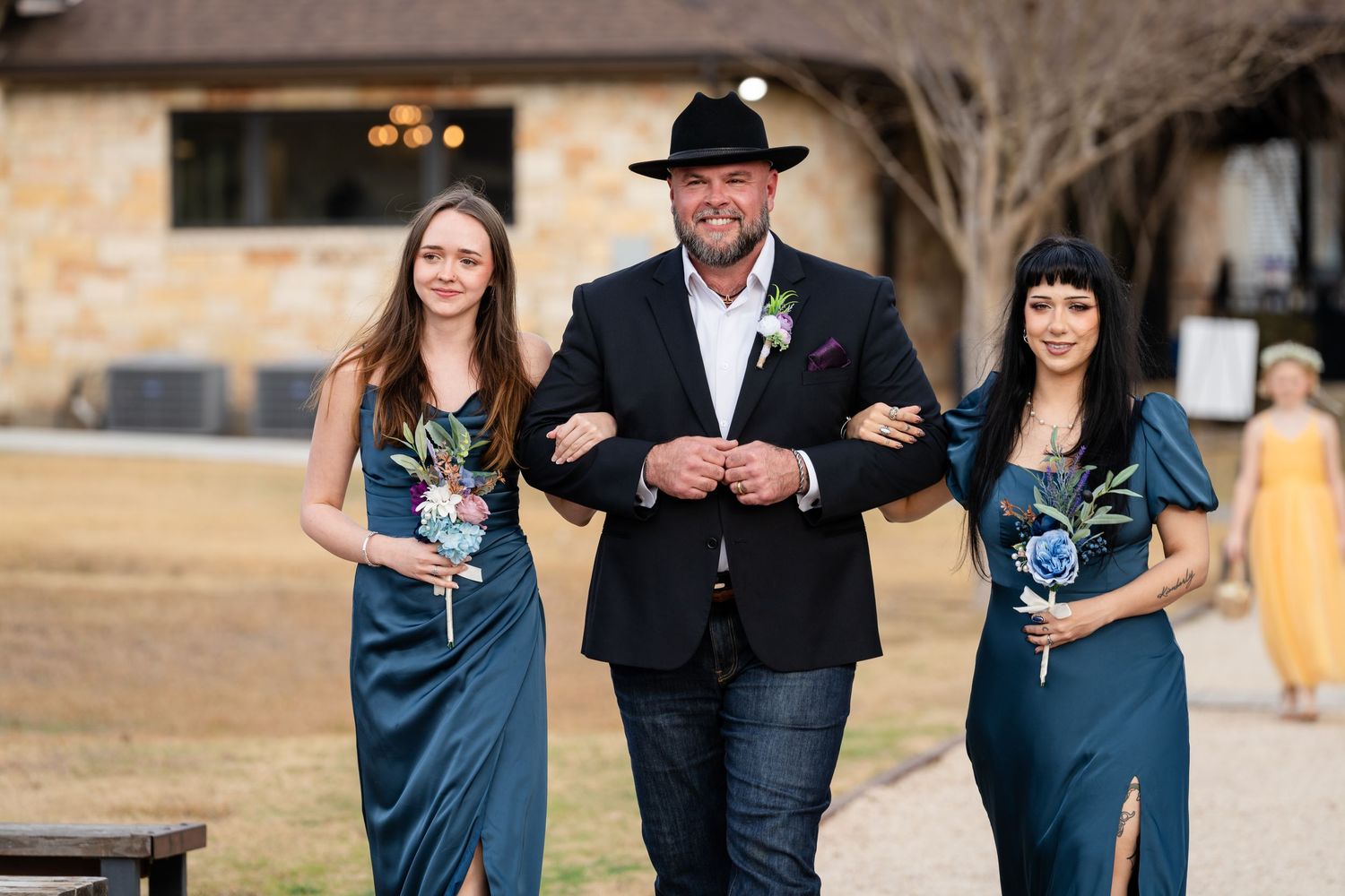 Man in black hat escorts two bridesmaids in teal gowns down wedding aisle at rustic stone venue.