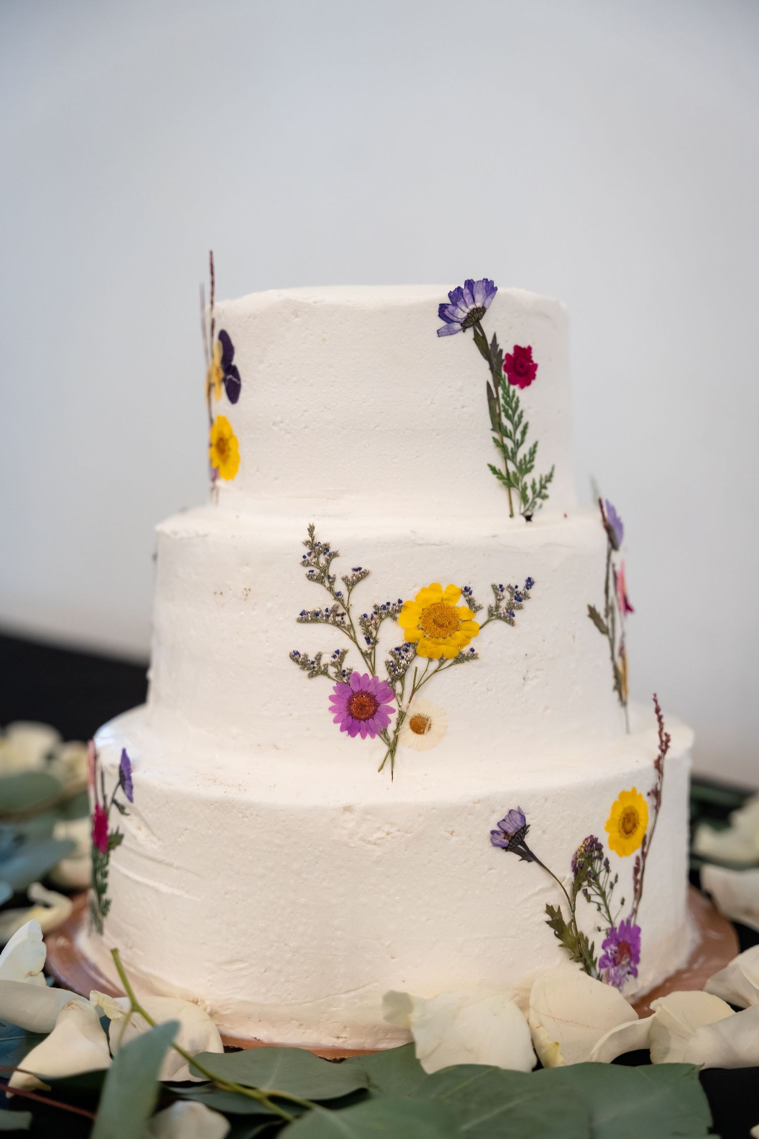 Three-tier white wedding cake decorated with colorful pressed wildflowers including yellow, purple, and pink blooms.
