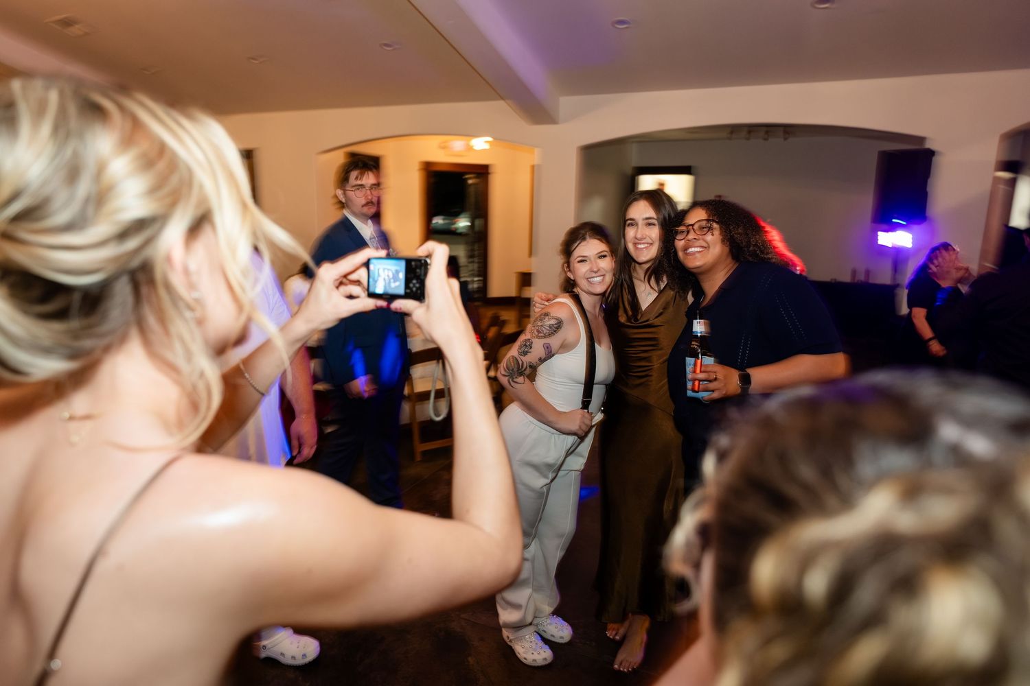 Group of young people posing for a photo at a party while a blonde woman photographs them with a smartphone.