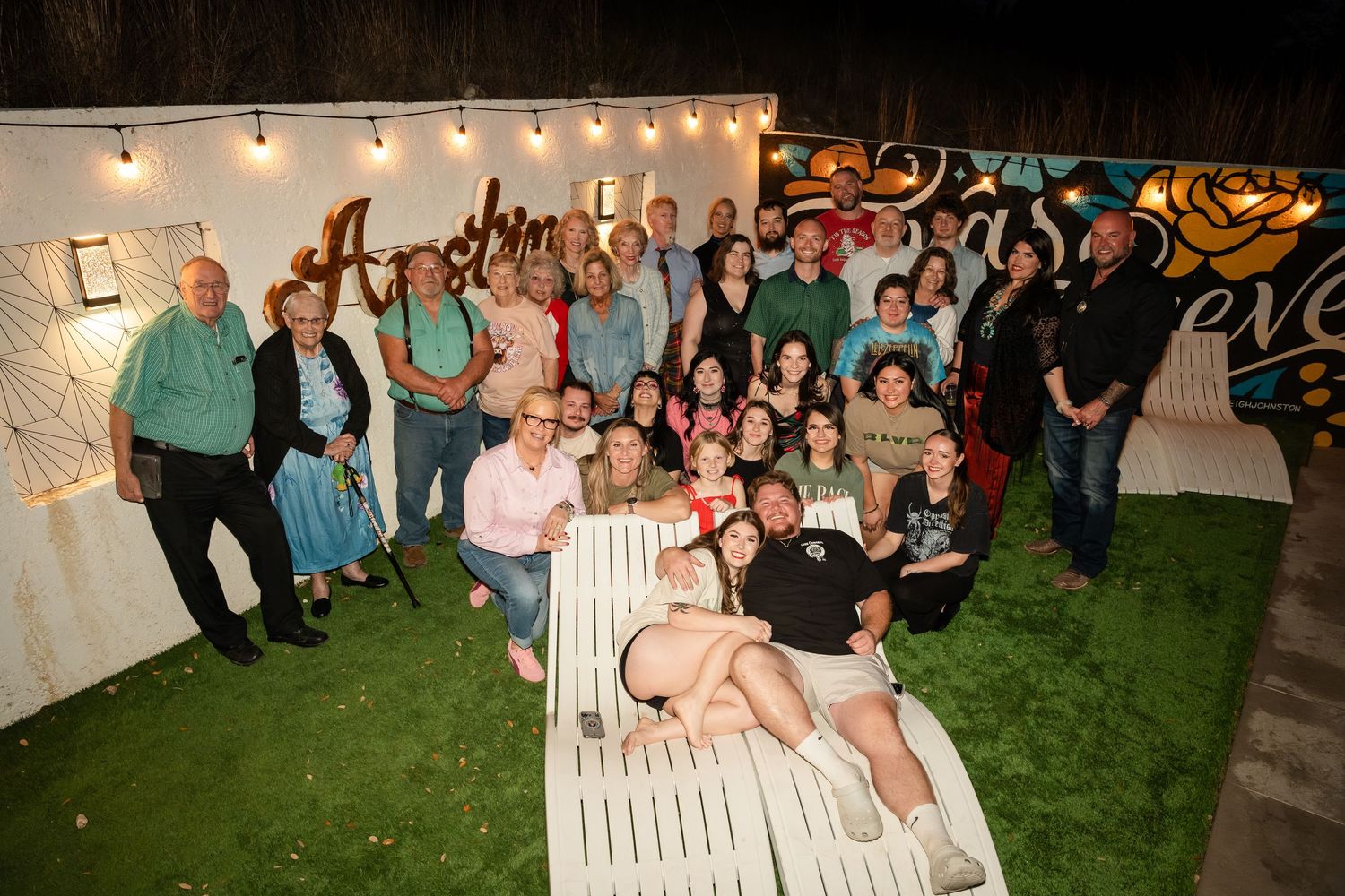 Large group of people posing together outdoors at night near decorative string lights and colorful mural backdrop.
