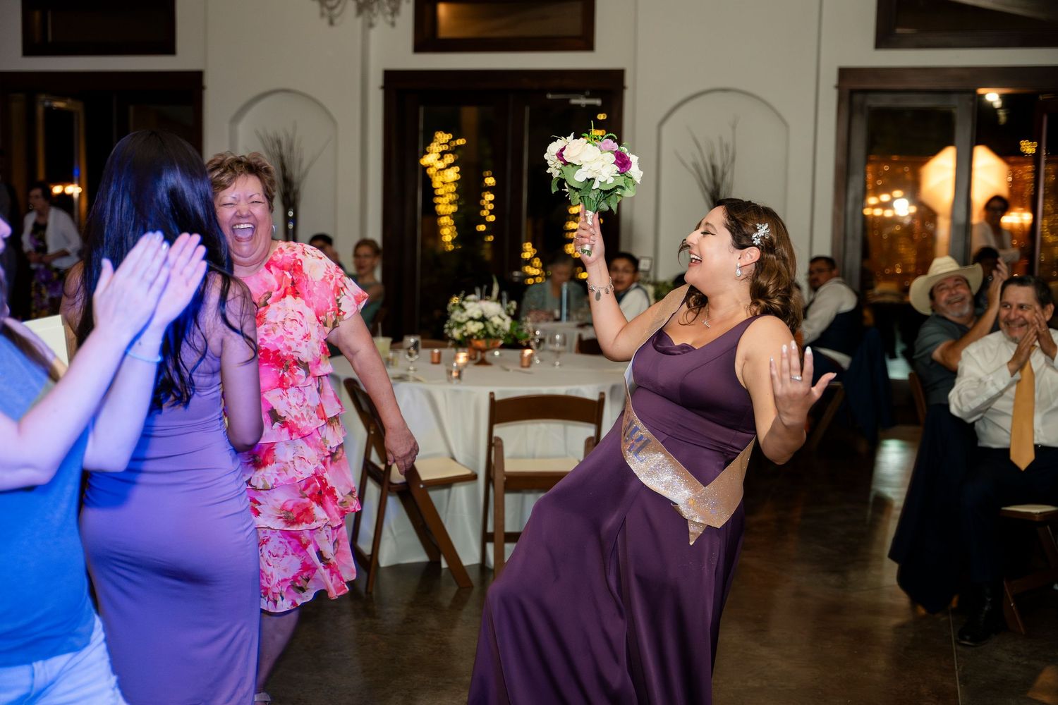 Guests dancing and celebrating with bouquet toss at indoor wedding reception with warm ambient lighting.