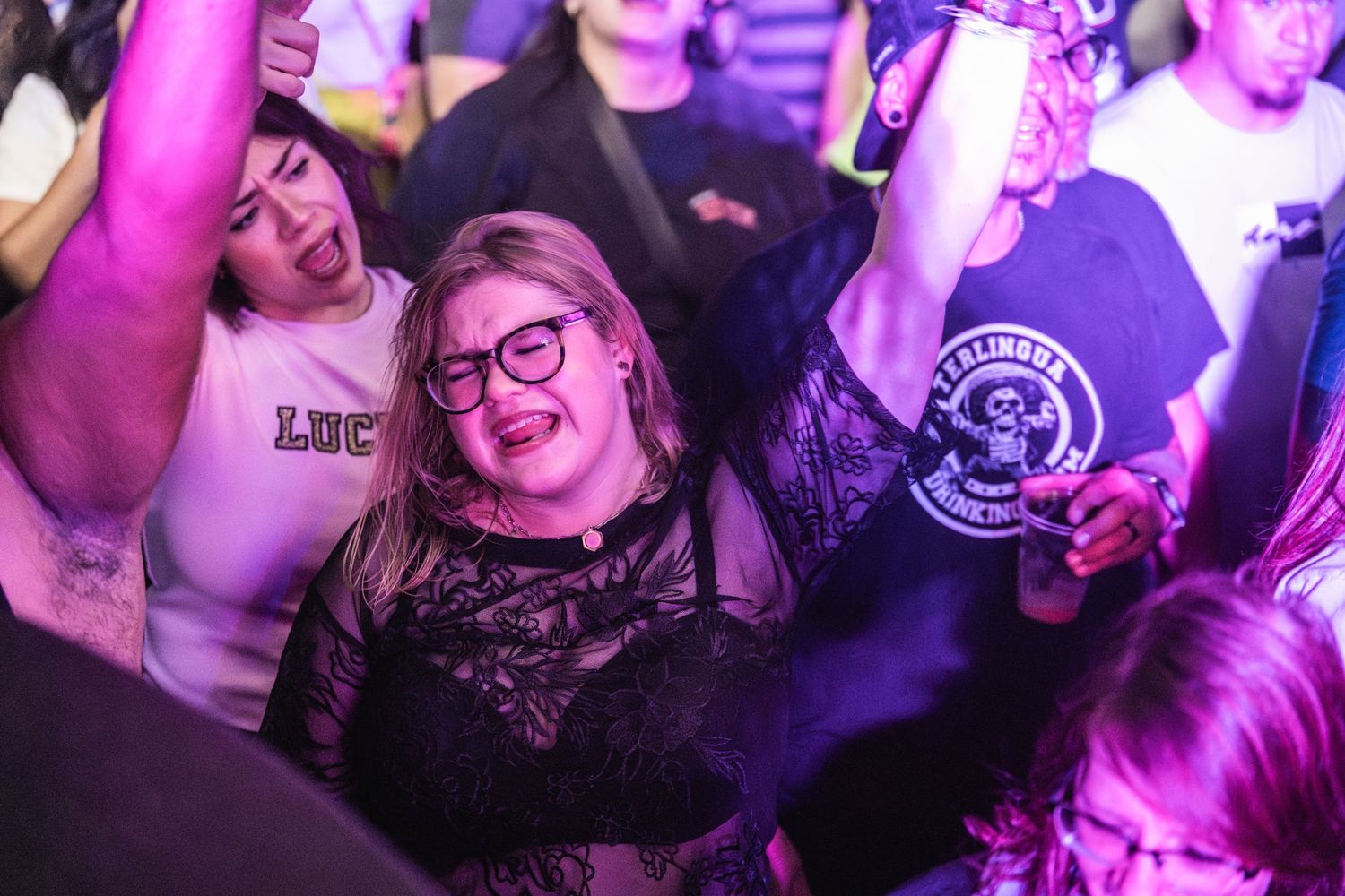 Concert attendees dance and celebrate with raised drinks under purple lighting at a music event.