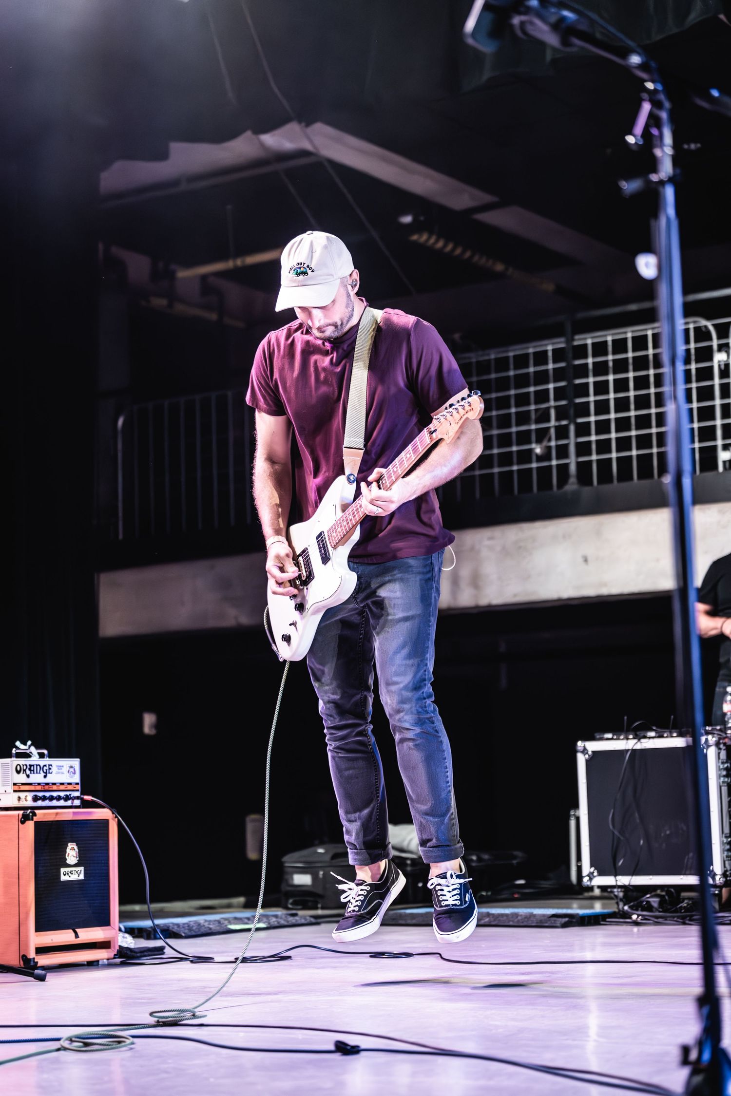 A musician plays electric guitar on stage wearing a burgundy shirt and white cap under stage lighting.