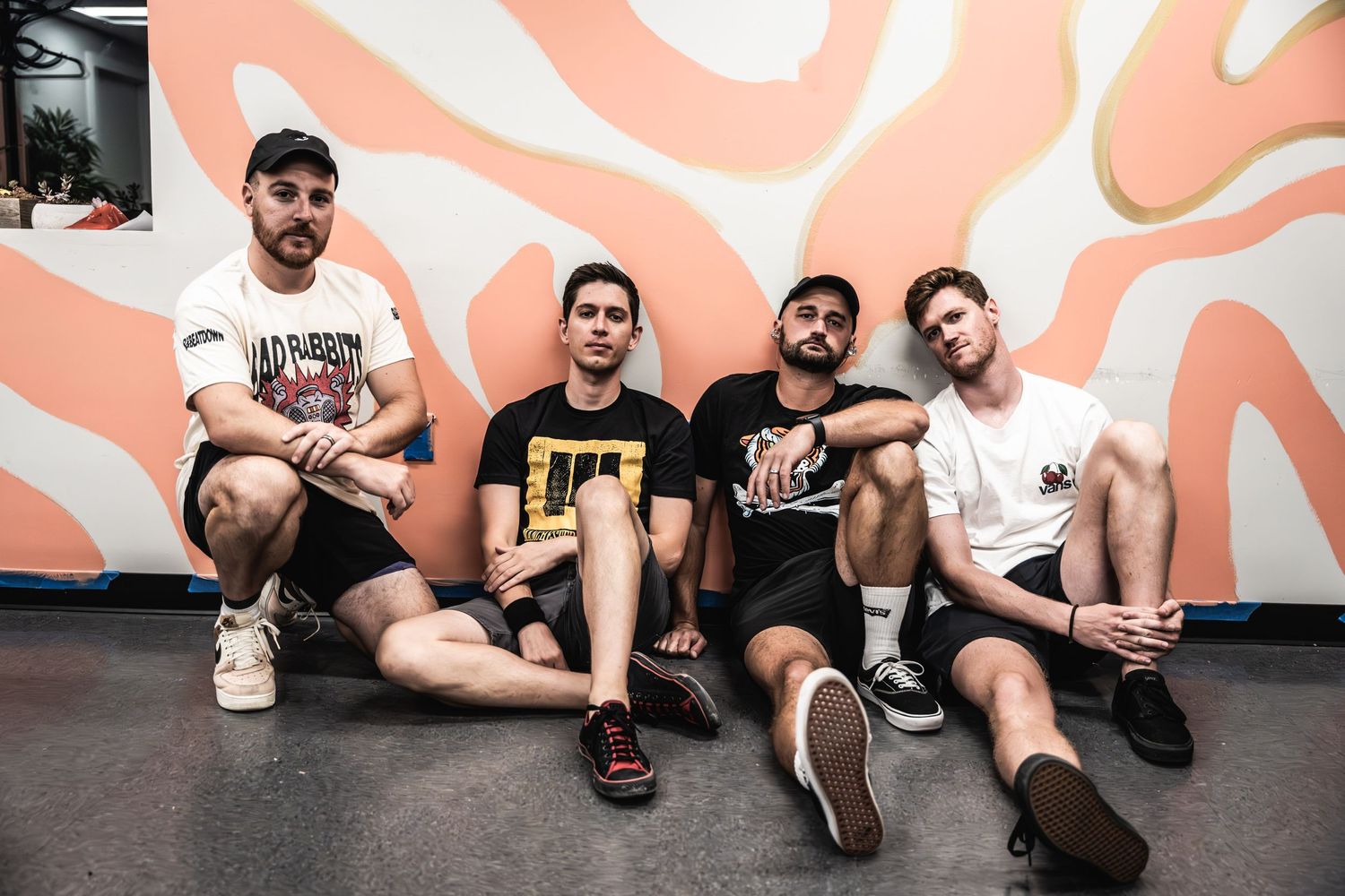 Musicians sit against a coral-colored wall with abstract white patterns.