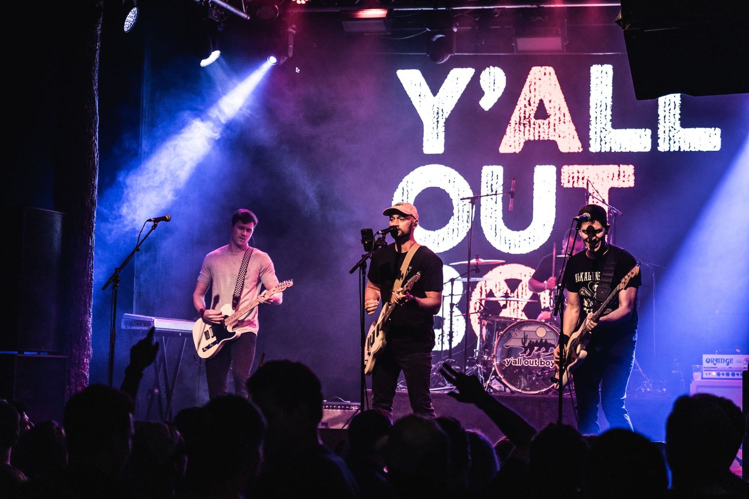 Band performing under Y'ALL OUT sign with blue stage lighting and silhouetted audience.