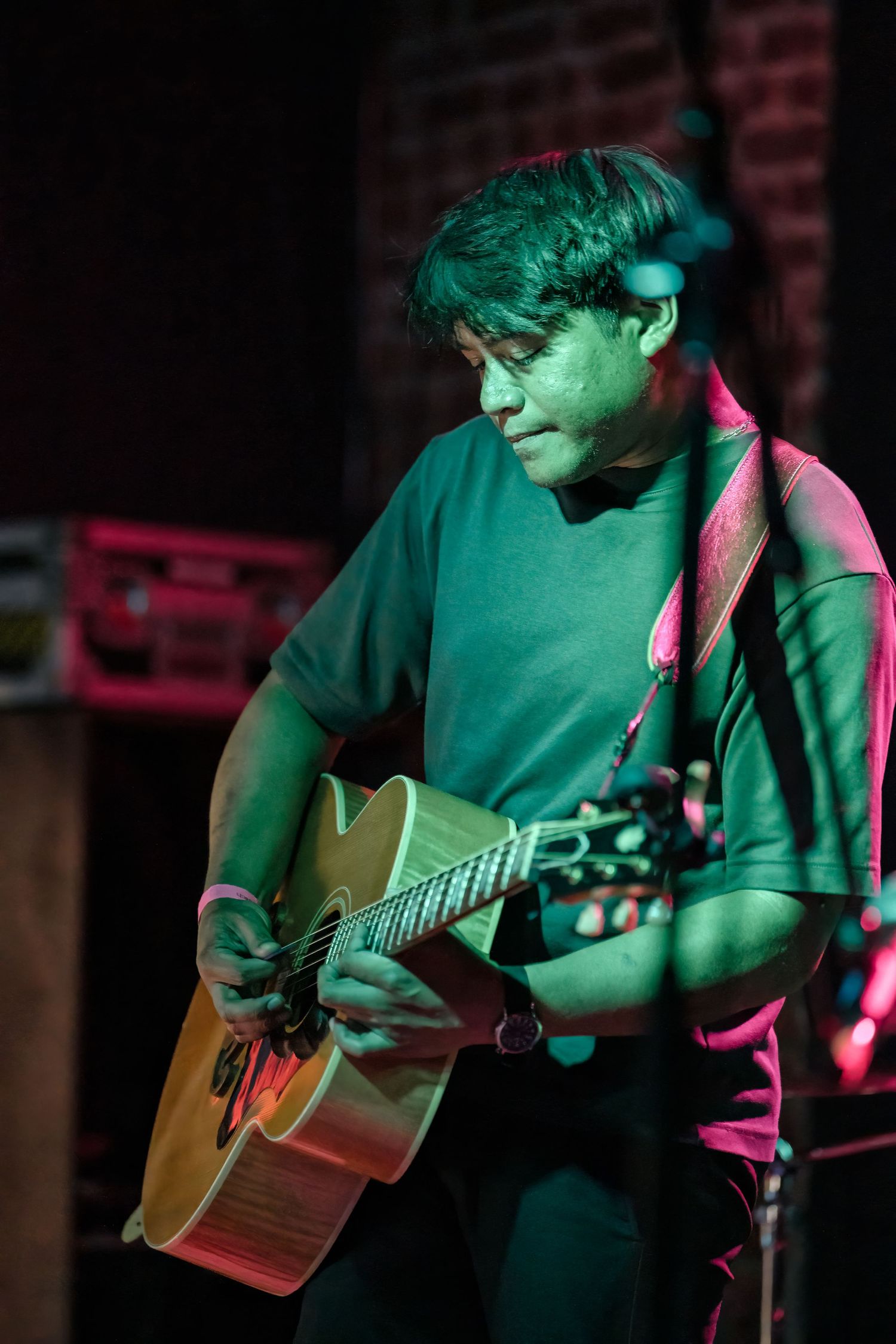 Musician strums acoustic guitar bathed in dramatic green and pink stage lighting at intimate venue.