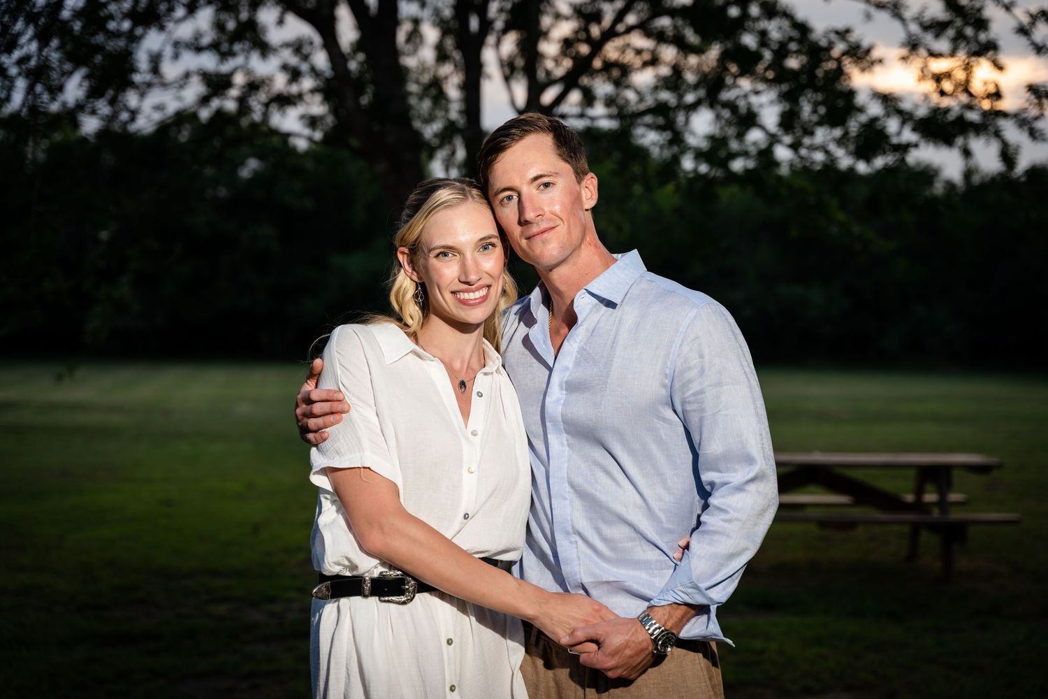Elegant couple poses together at dusk in a park setting wearing coordinated casual attire.