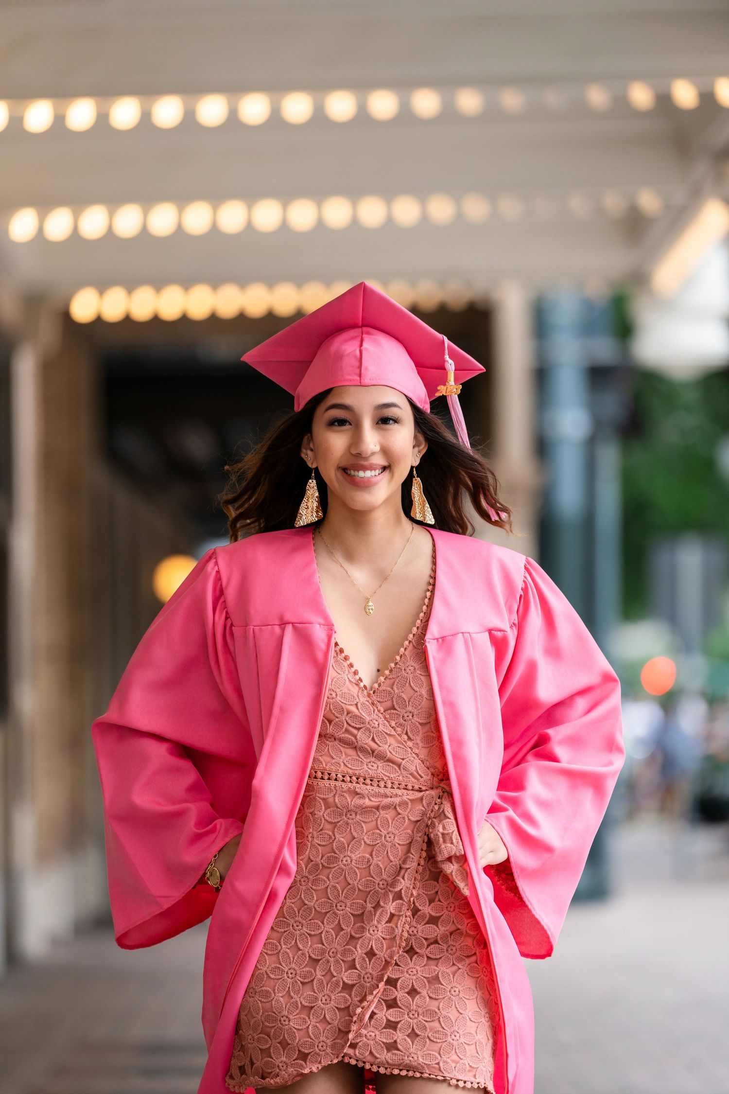 Graduate in bright pink cap and gown poses under string lights.