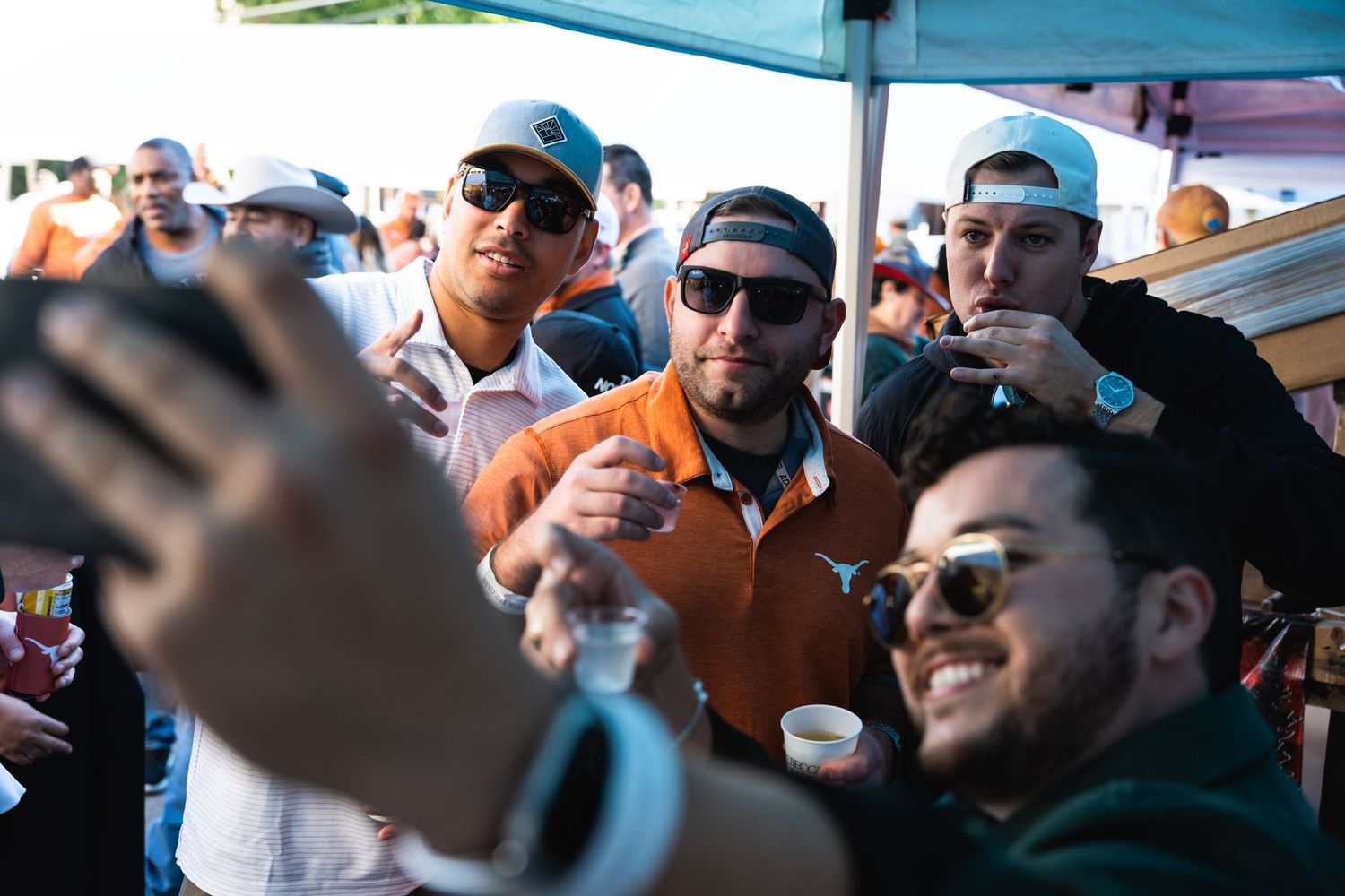 Enthusiastic crowd enjoying drinks under a blue canopy tent at an outdoor festival.