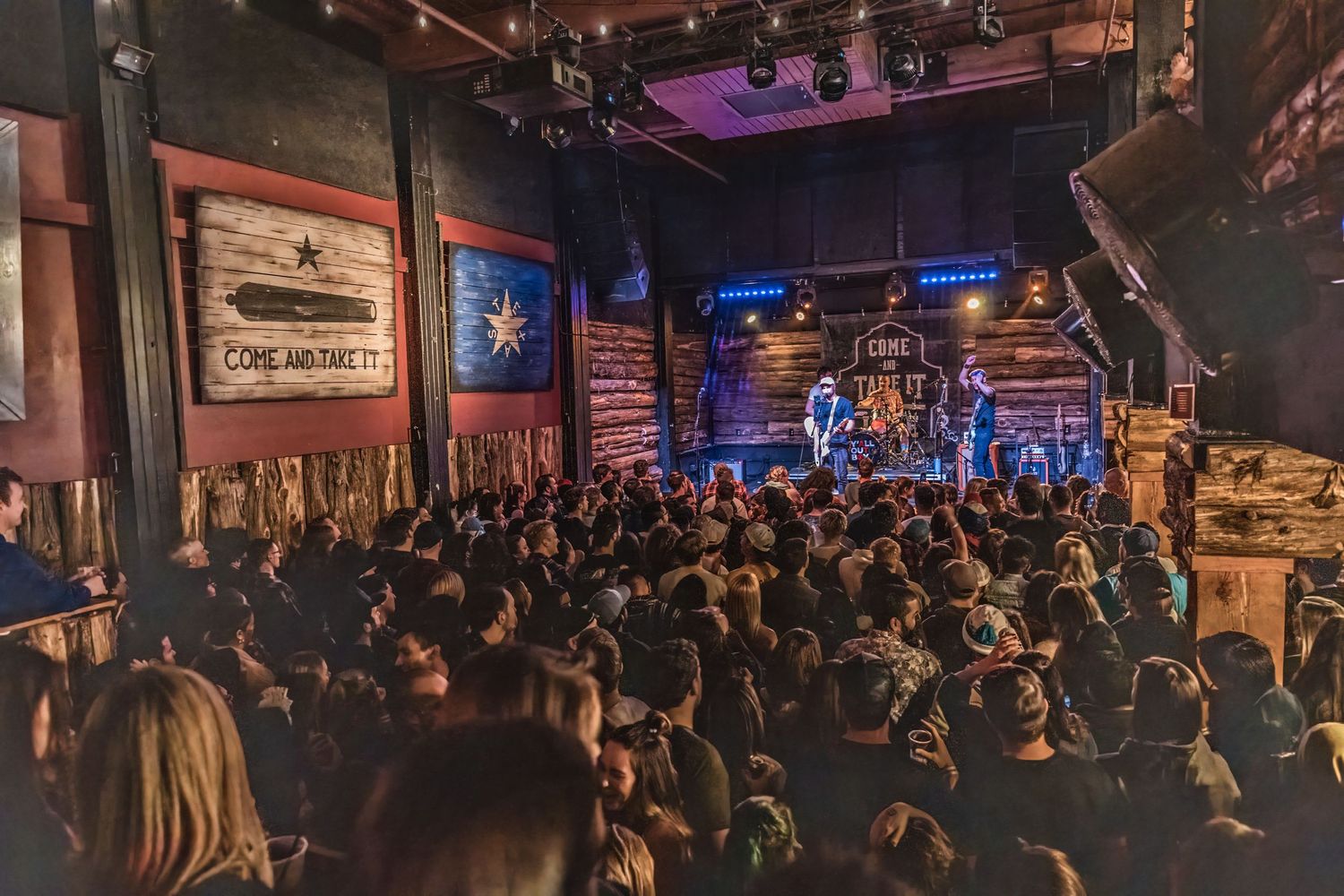 Packed concert venue with enthusiastic crowd watching performers on illuminated stage.