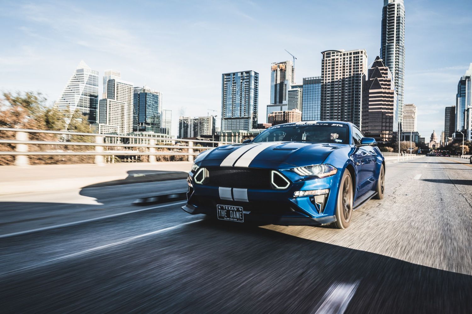 Blue Ford Mustang GT with racing stripes cruising past Austin skyline on elevated highway.