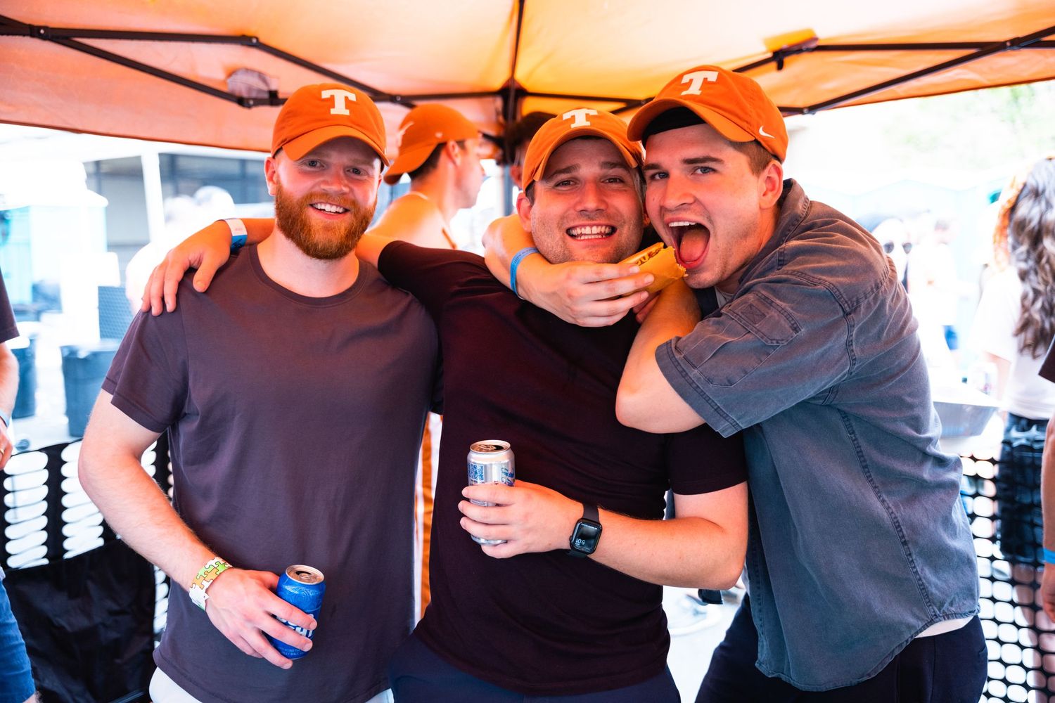 Three friends in orange baseball caps enjoying drinks together under an orange tent at an outdoor event.