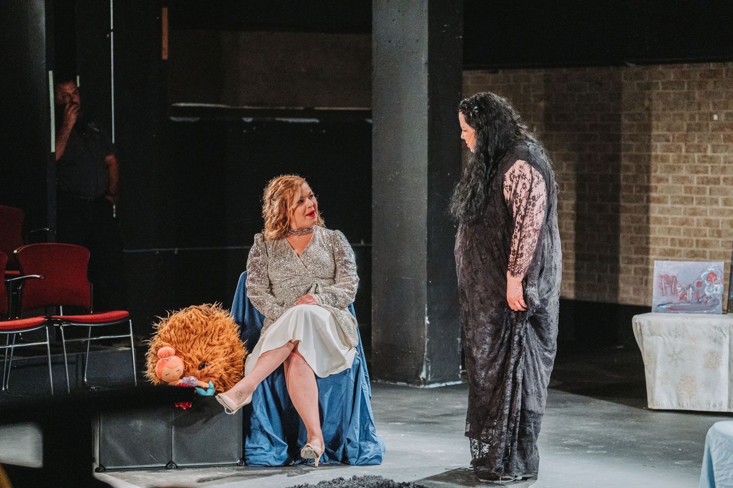 Two actors perform a dramatic scene on stage with one seated holding a prop.