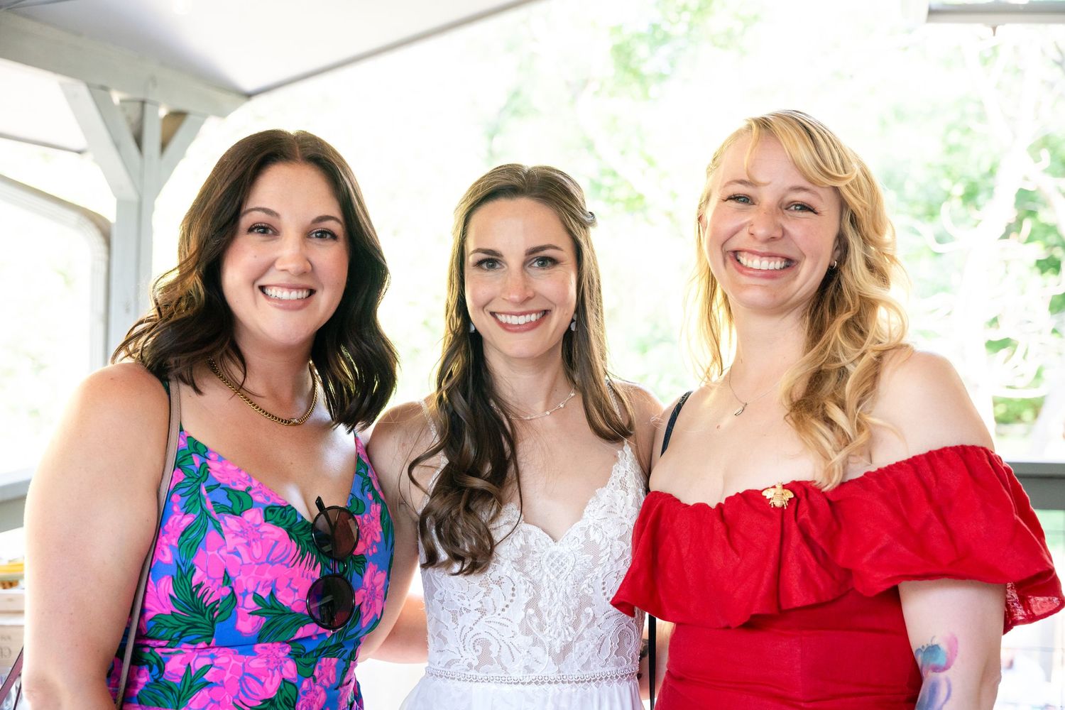 Three women in colorful dresses and white pose together at wedding celebration.