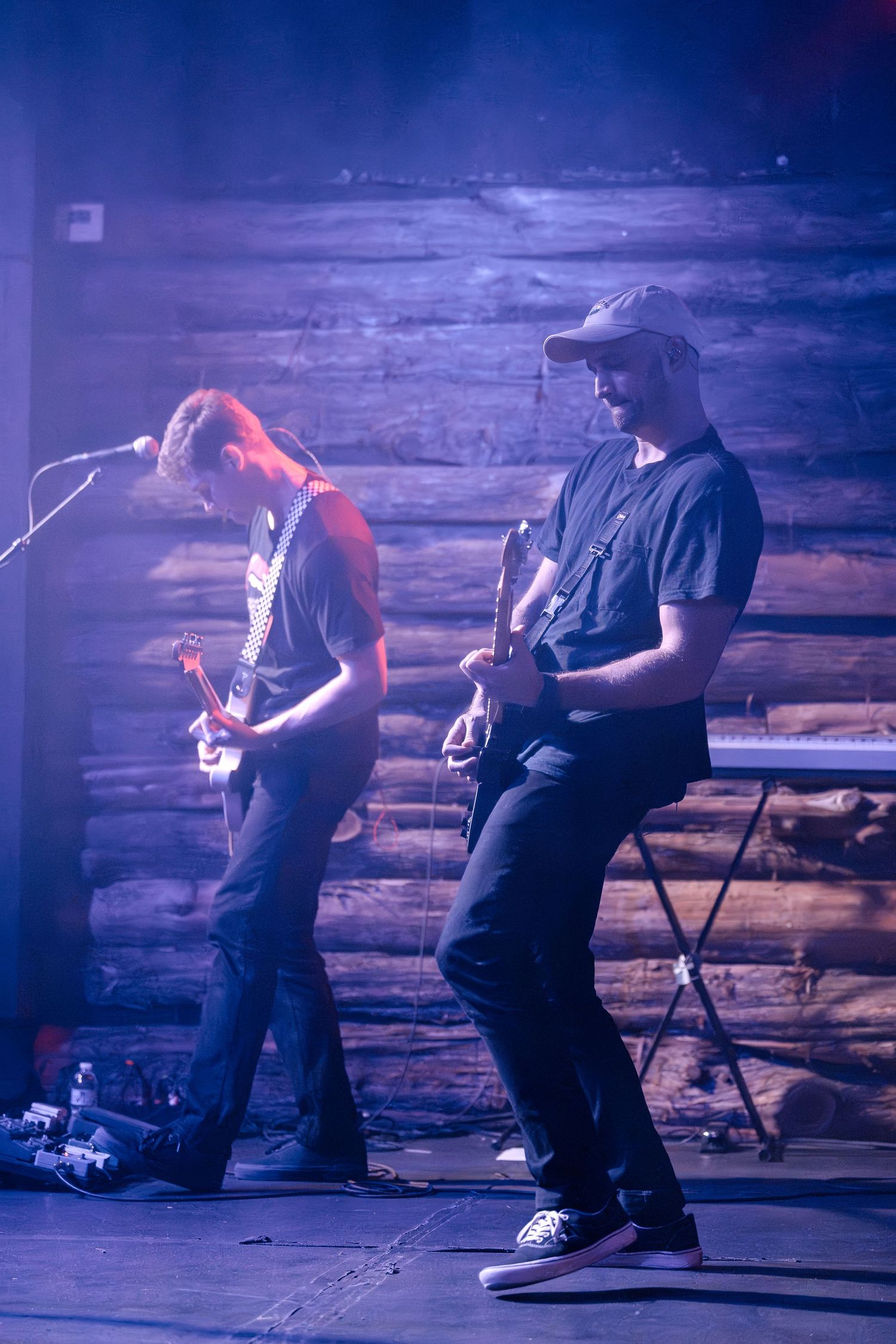 Two guitarists perform on stage with wooden wall backdrop and moody purple lighting.