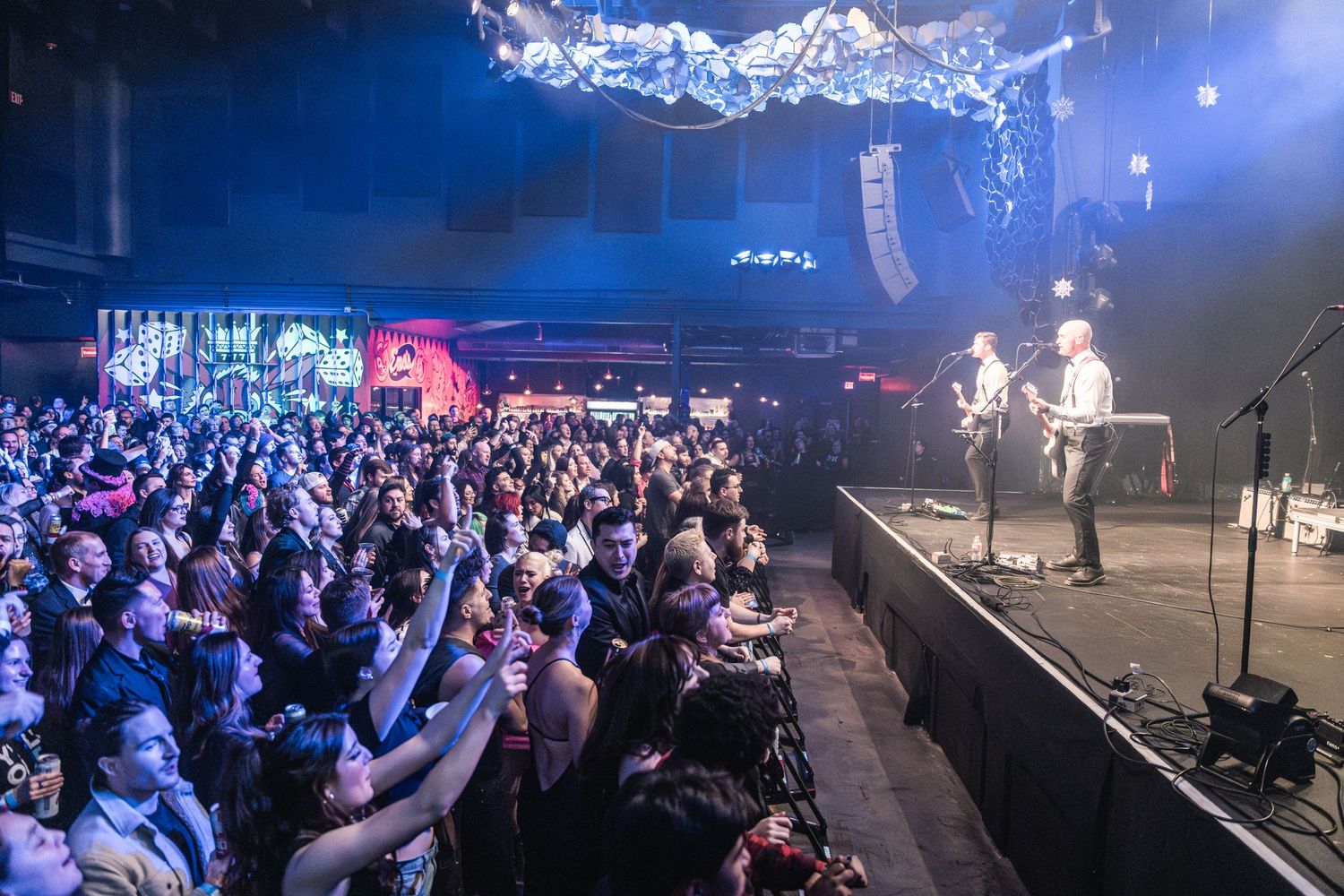 Wide shot of packed concert venue with crowd facing illuminated stage and band performing.