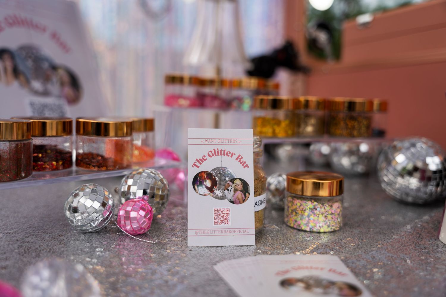 Small glass jars filled with gold glitter and sparkles arranged with pink and silver disco balls on a glittery surface.
